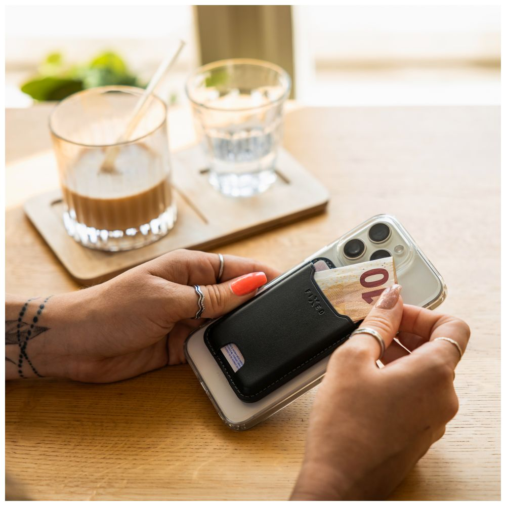 Hände halten ein Telefon mit einer Geldbörsenhülle und einer Banknote. Kaffee, Wasser und Holztablett im Hintergrund.