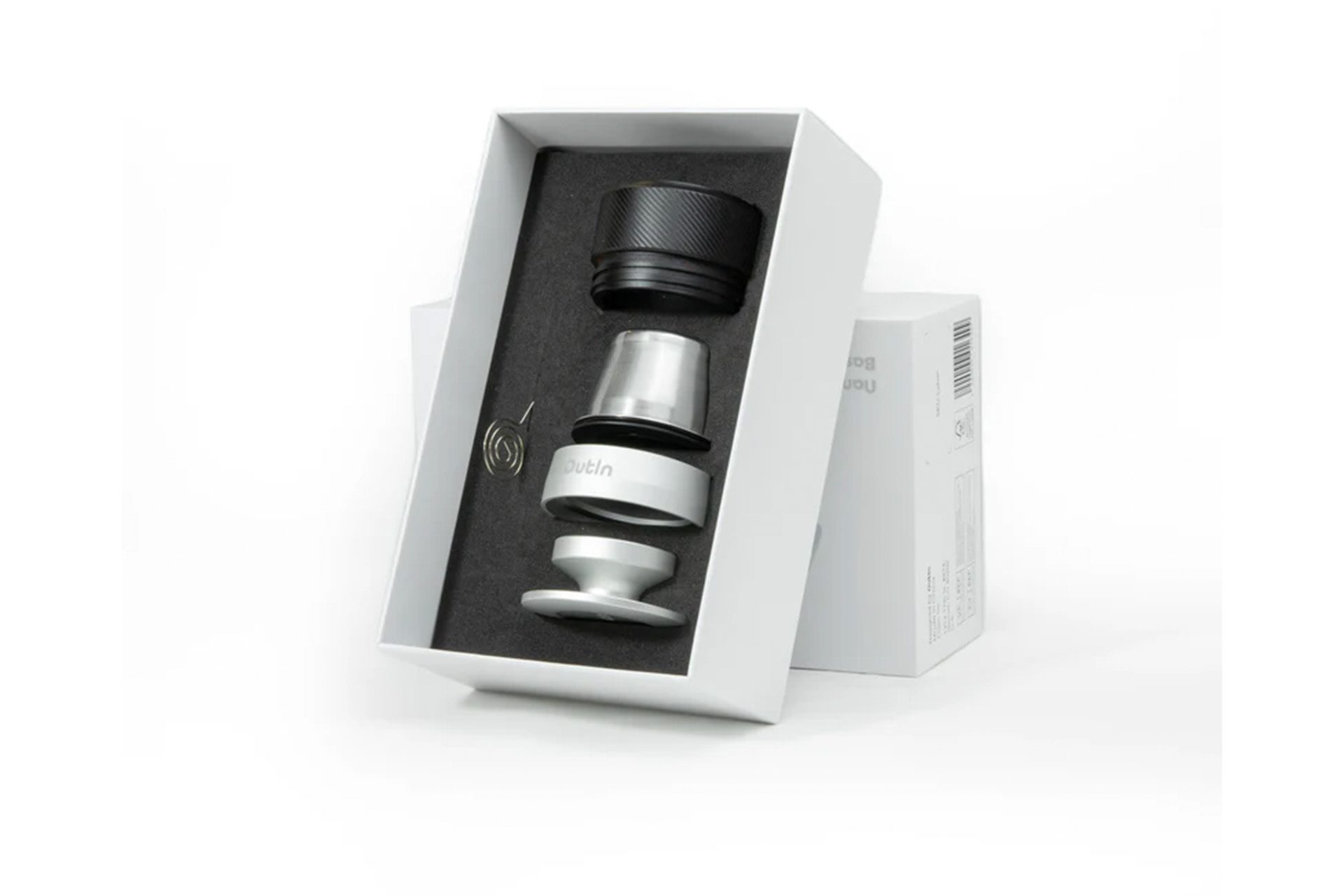 Otwarta biała skrzynka z częściami w środku: czarna góra, srebrna środkowa część, biała podstawa i sprężyna.