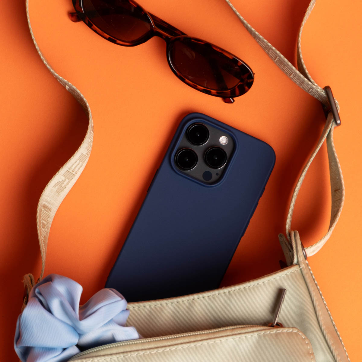 Sonnenbrille und Telefon in einer beigen Tasche auf orangefarbenem Hintergrund. Ein blaues Scrunchie ist sichtbar.