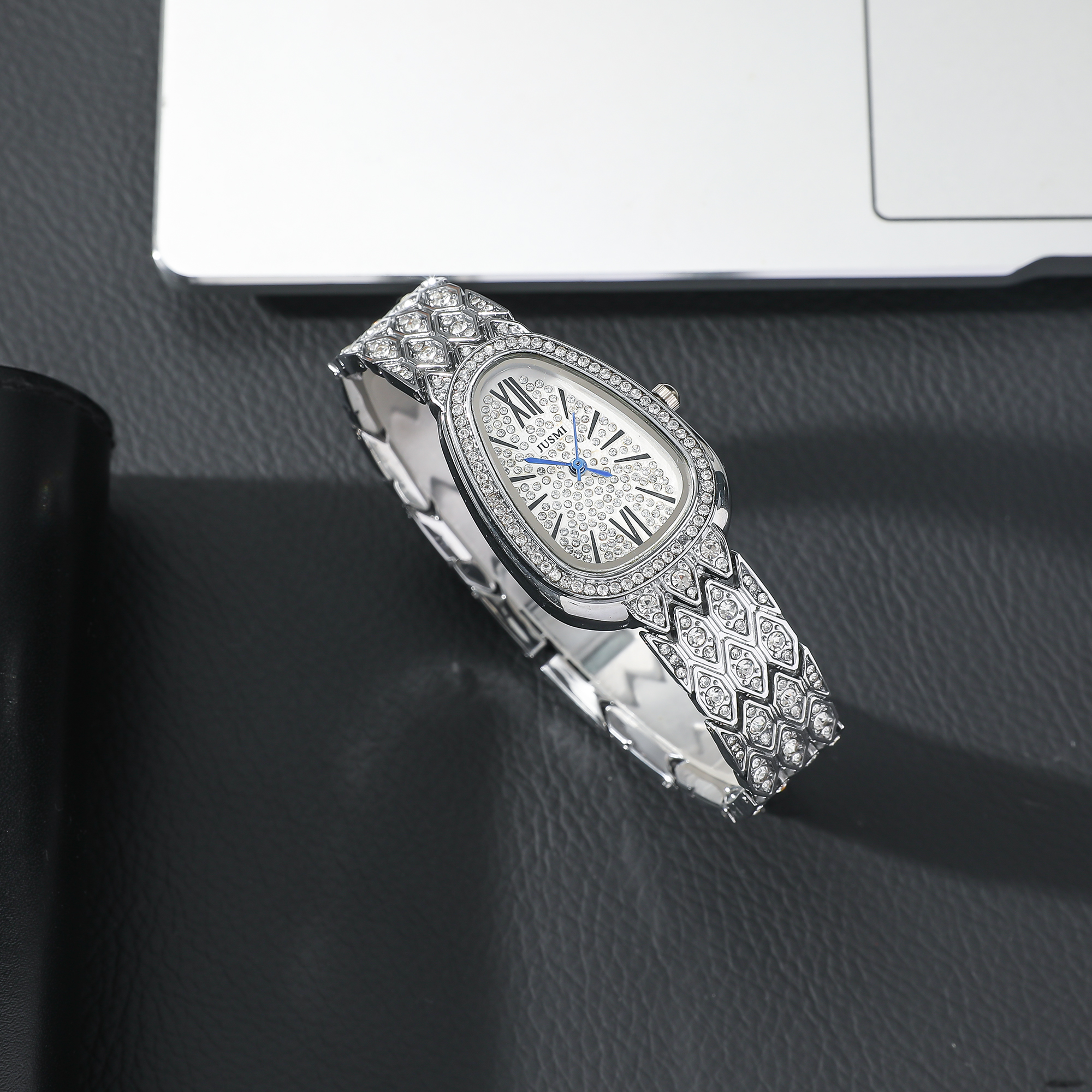 Silberne Uhr mit diamantartigem Muster auf schwarzem Untergrund. Laptop im Hintergrund sichtbar.