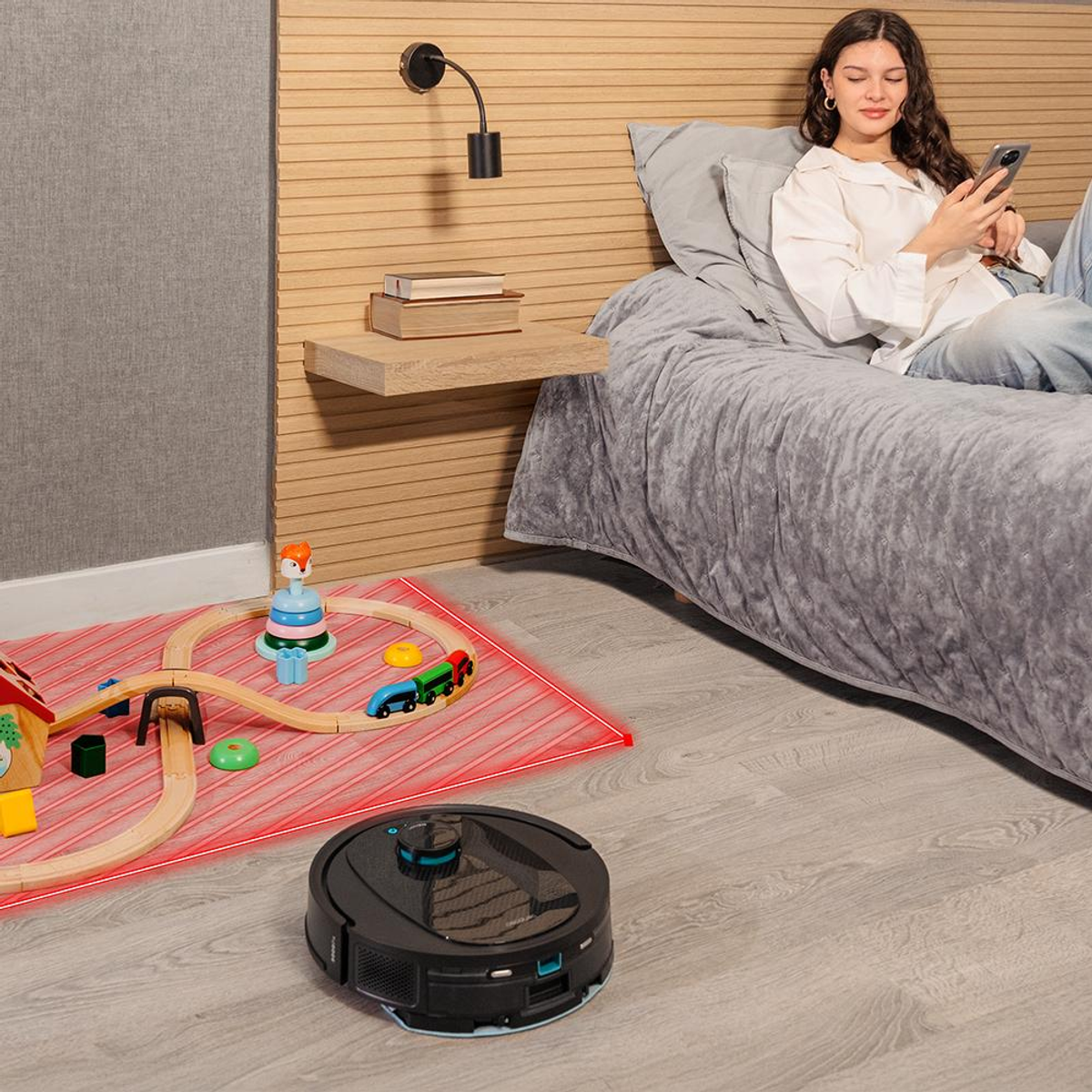 Sala de estar con mujer en cama, robot aspirador y tren de juguete. Línea roja indica zona de limpieza.