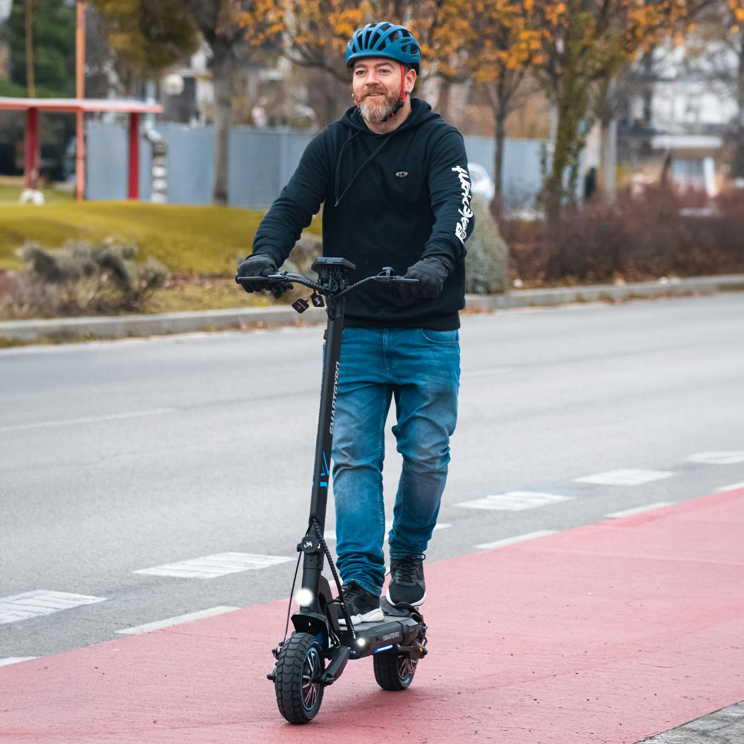 Un hombre con una sudadera con capucha negra y jeans montando un patinete eléctrico en una carretera roja.