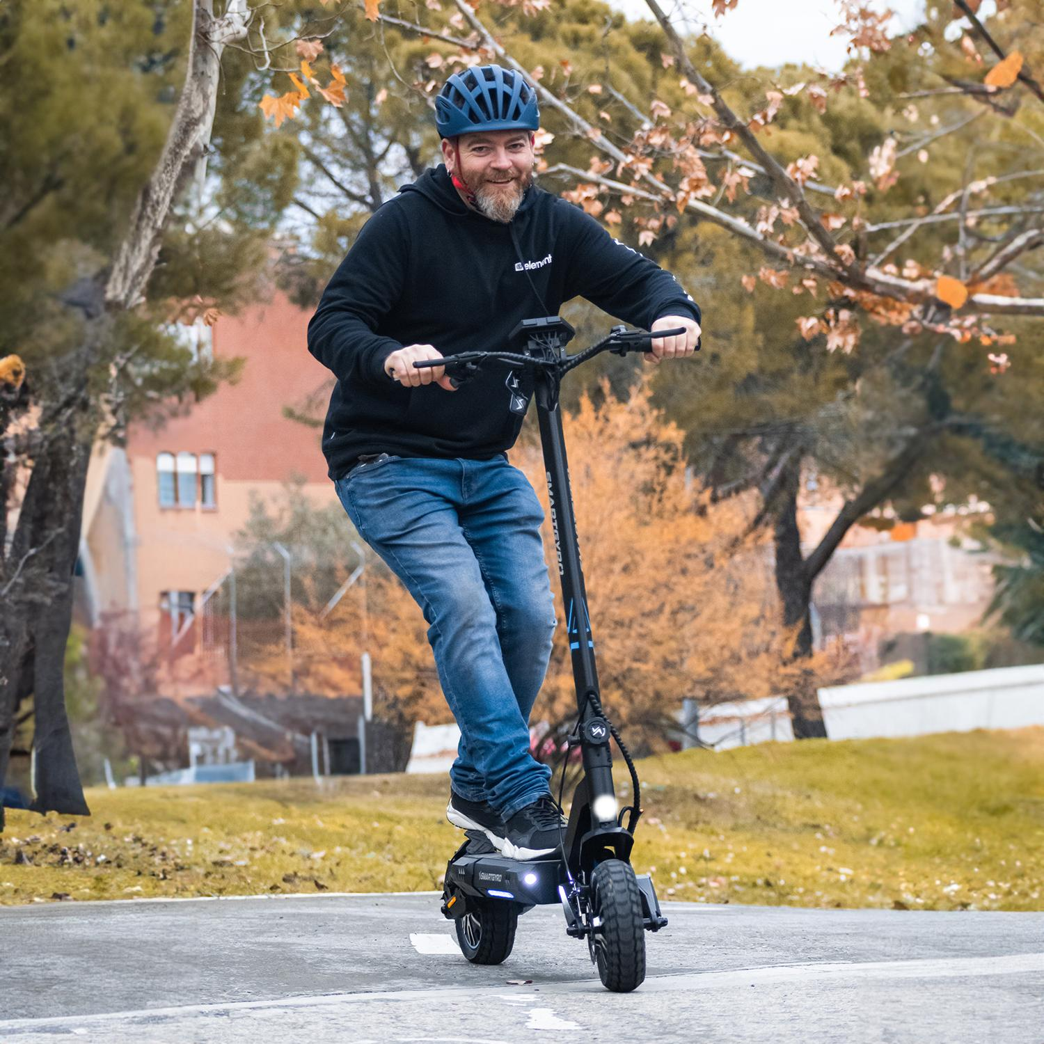 Hombre montando un patinete eléctrico en una carretera con árboles al fondo. Lleva un casco.