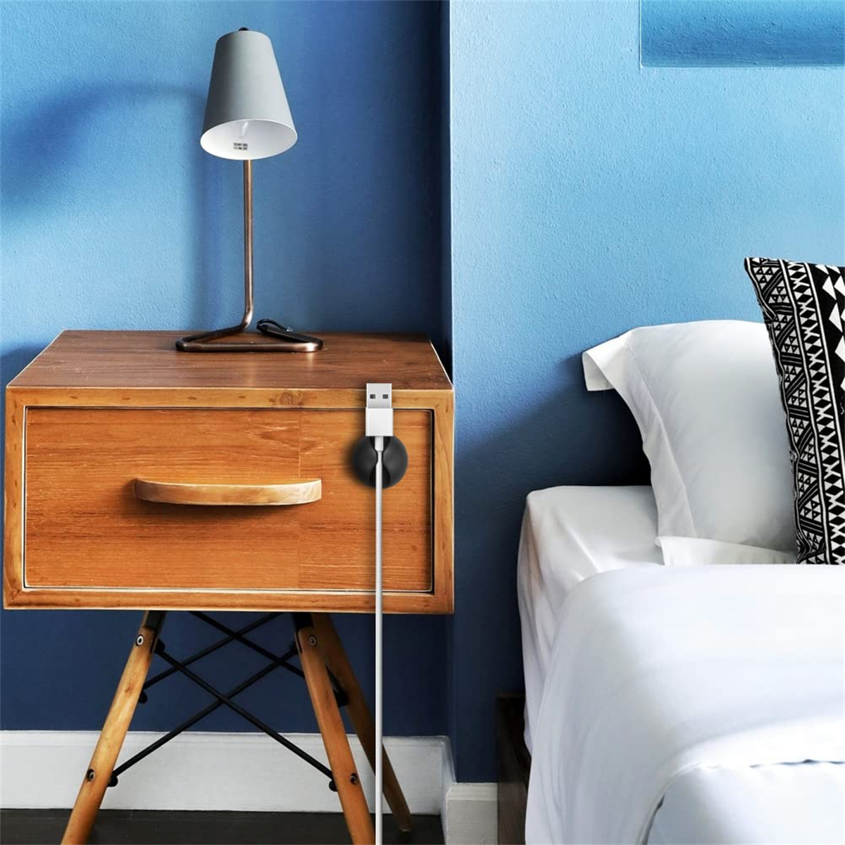 Nachttisch aus Holz mit Lampe. Weißes Bett und schwarz-weißes Kissen. Blaue Wand im Hintergrund.