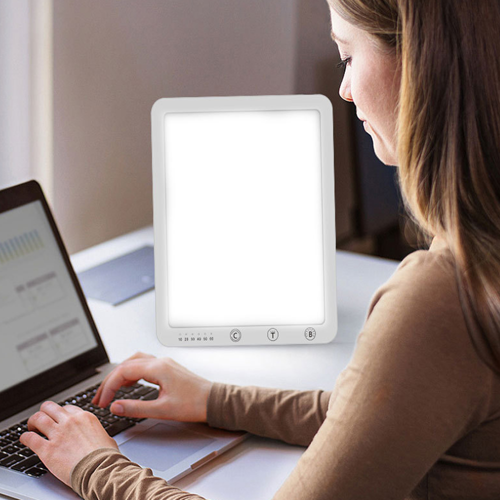 Frau benutzt eine weiße Lichttherapielampe. Laptop auf dem Schreibtisch. Helles weißes Licht an.