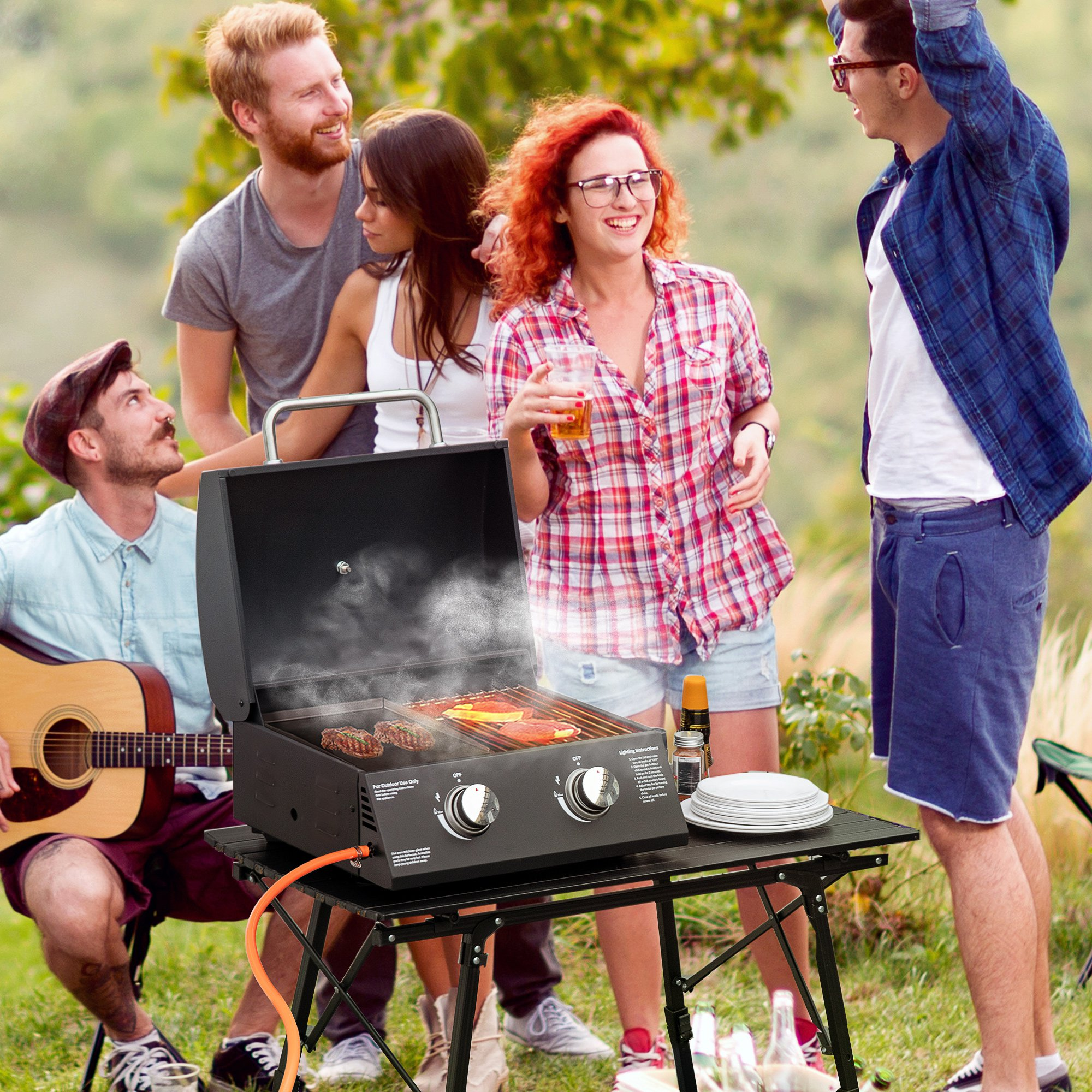Un grupo de personas en una barbacoa con una parrilla, mesa e instrumento musical en un entorno de parque.