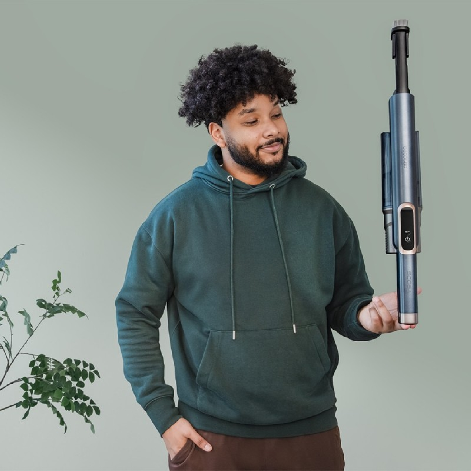 Hombre en sudadera con capucha verde sosteniendo una aspiradora. La mira sonriendo. Pared verde al fondo.