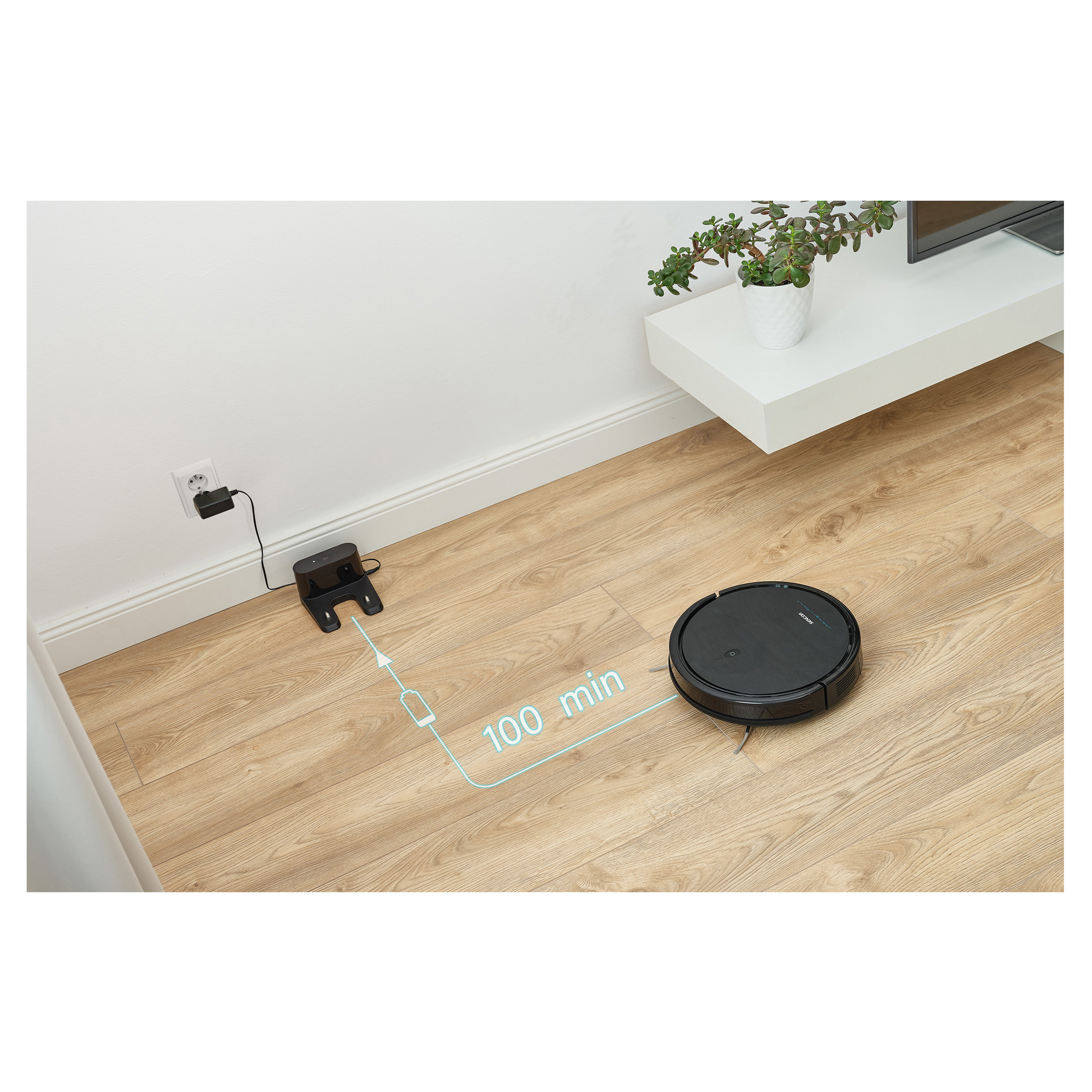 Black robot vacuum cleaner on wooden floor, charging, with a white wall and furniture in the background.