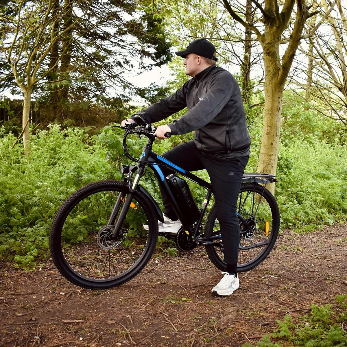 Osoba jadąca czarno-niebieskim rowerem elektrycznym na ścieżce otoczonej drzewami i zielenią.