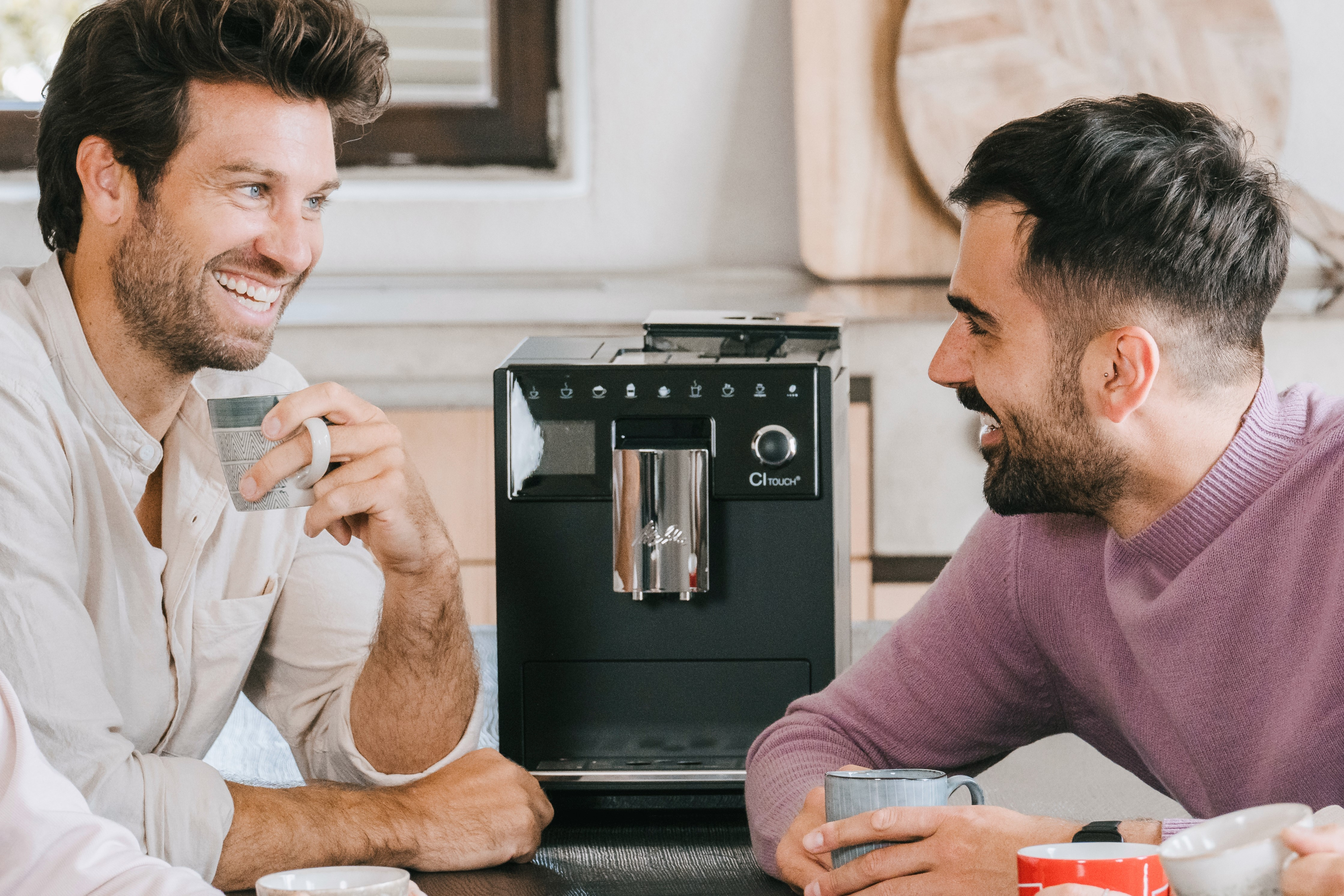 Dwóch uśmiechniętych mężczyzn trzymających kubki przed ekspresem do kawy w jasnej kuchni.