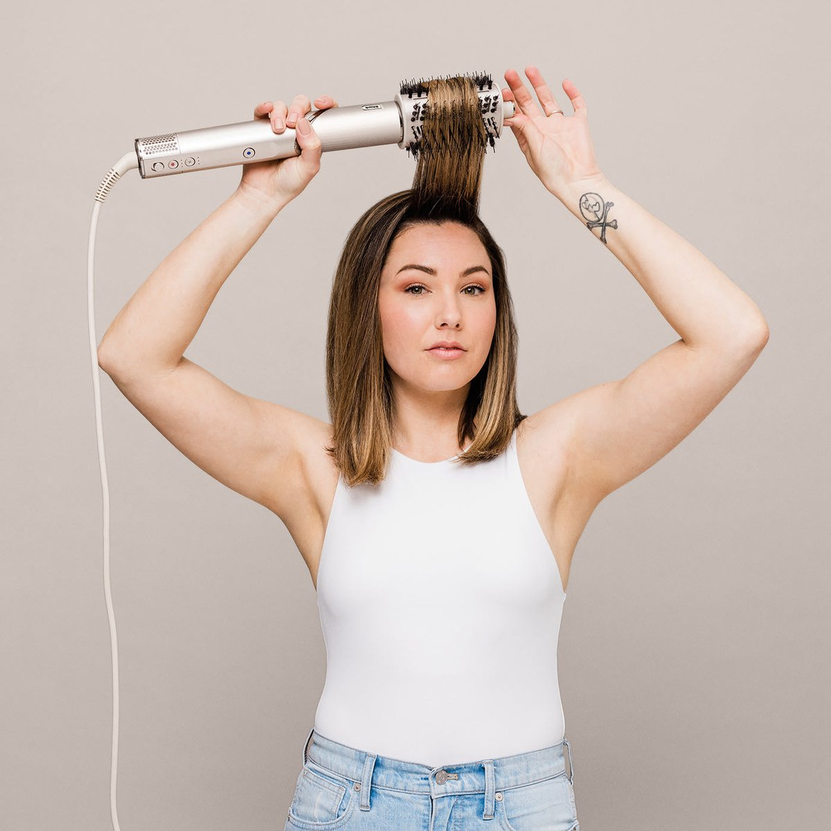Kobieta stylizuje włosy srebrnym urządzeniem do stylizacji, podniesione ramiona. Tatuaż na ramieniu.