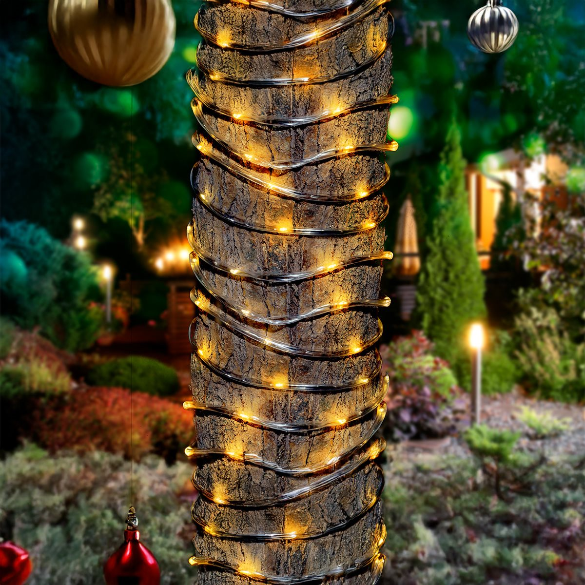 A tree trunk wrapped with yellow fairy lights, in a garden setting with ornaments.