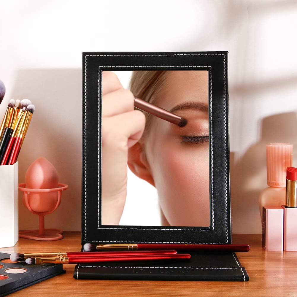 Eine Frau blickt in einen schwarz gerahmten Spiegel auf einem Holztisch. Make-up-Pinsel und Produkte sind sichtbar.
