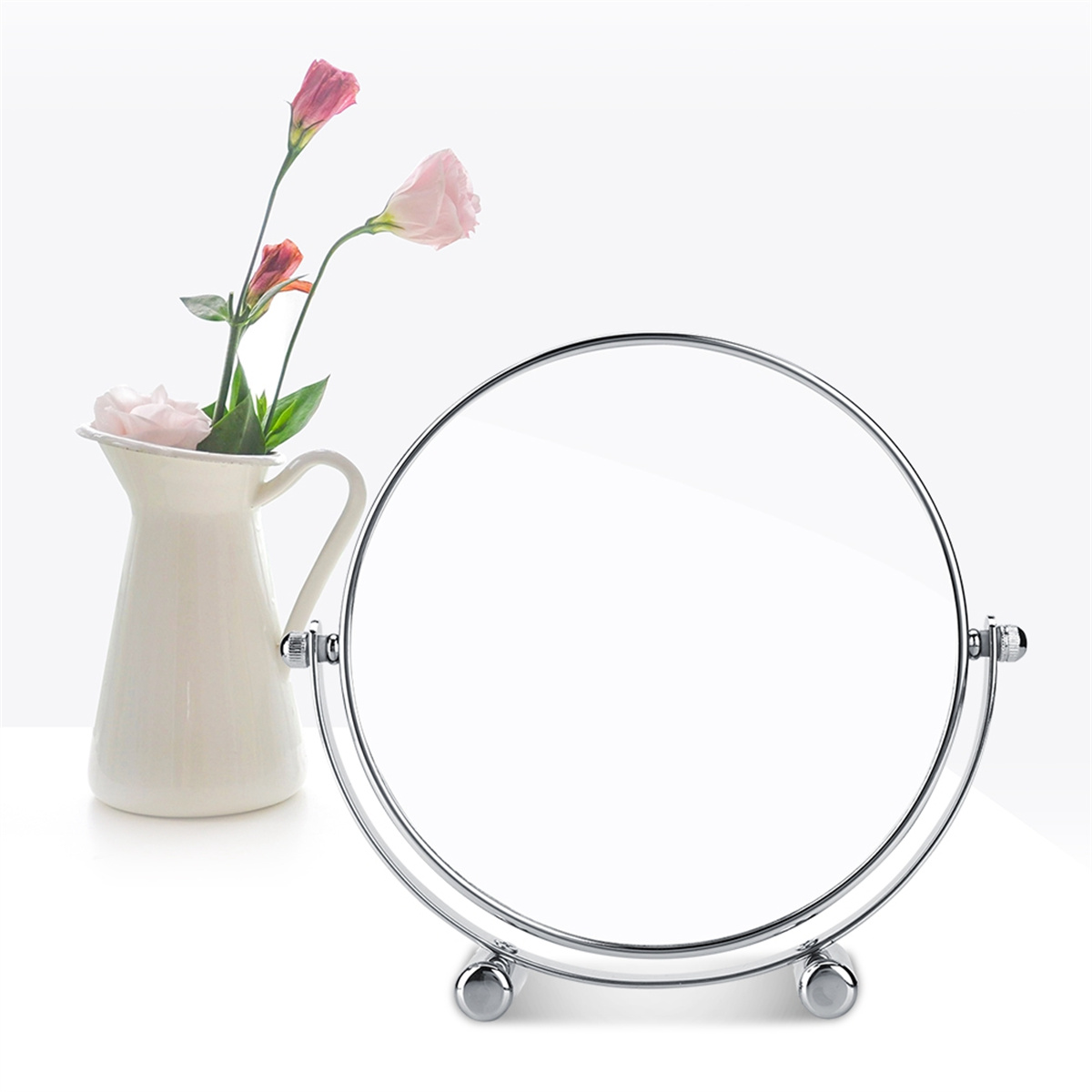 Ein runder Spiegel mit einer Vase mit rosa Blumen links, auf weißer Oberfläche.