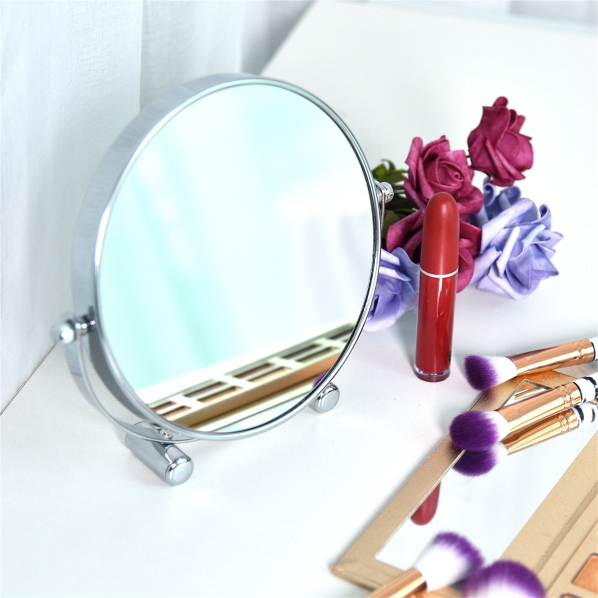 Runder Spiegel auf weißem Tisch mit Lippenstift, Pinseln und Blumen im Hintergrund.