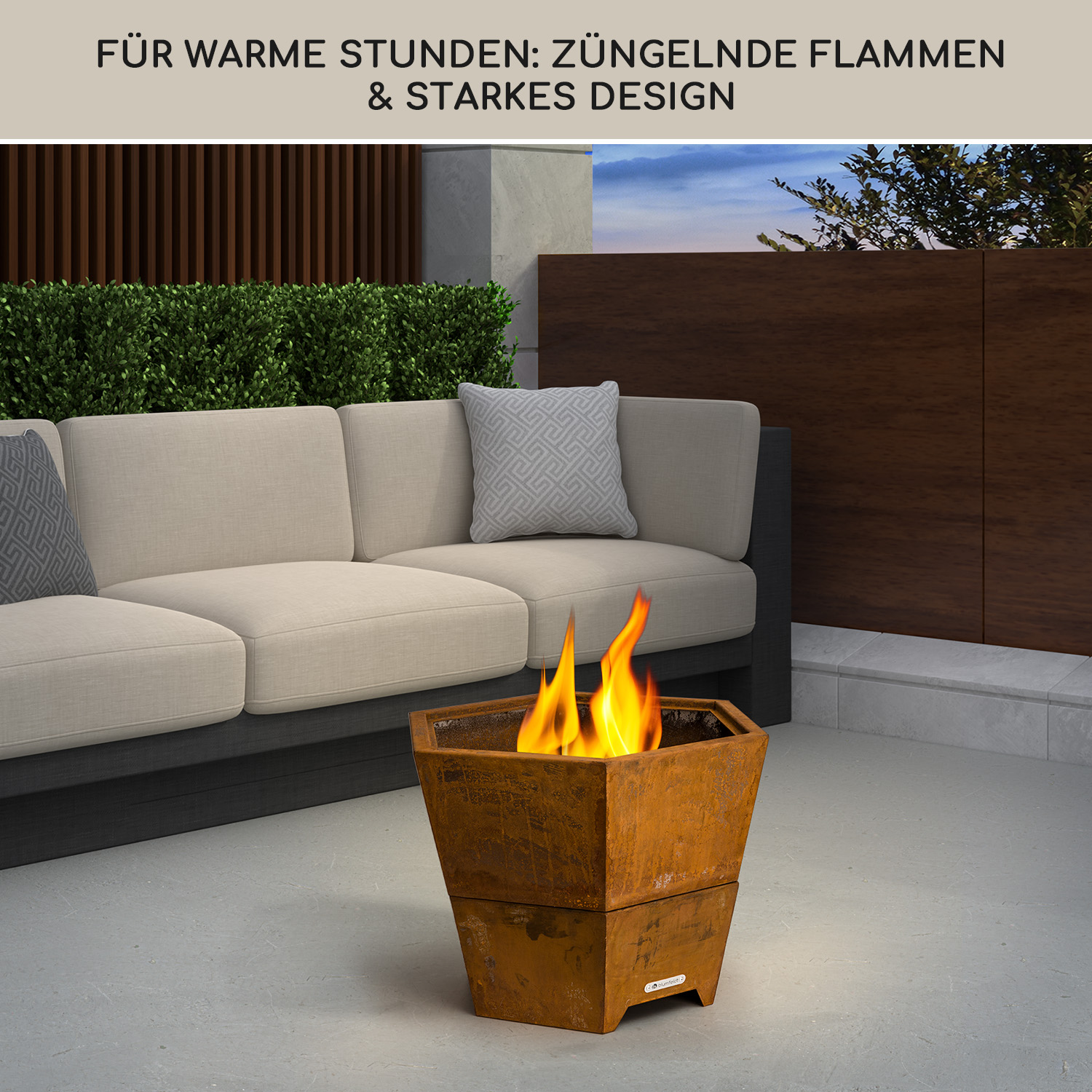 Eine brennende, rostig aussehende Feuerstelle mit Flammen auf einer Beton-Terrasse mit Sofa und Pflanzen.