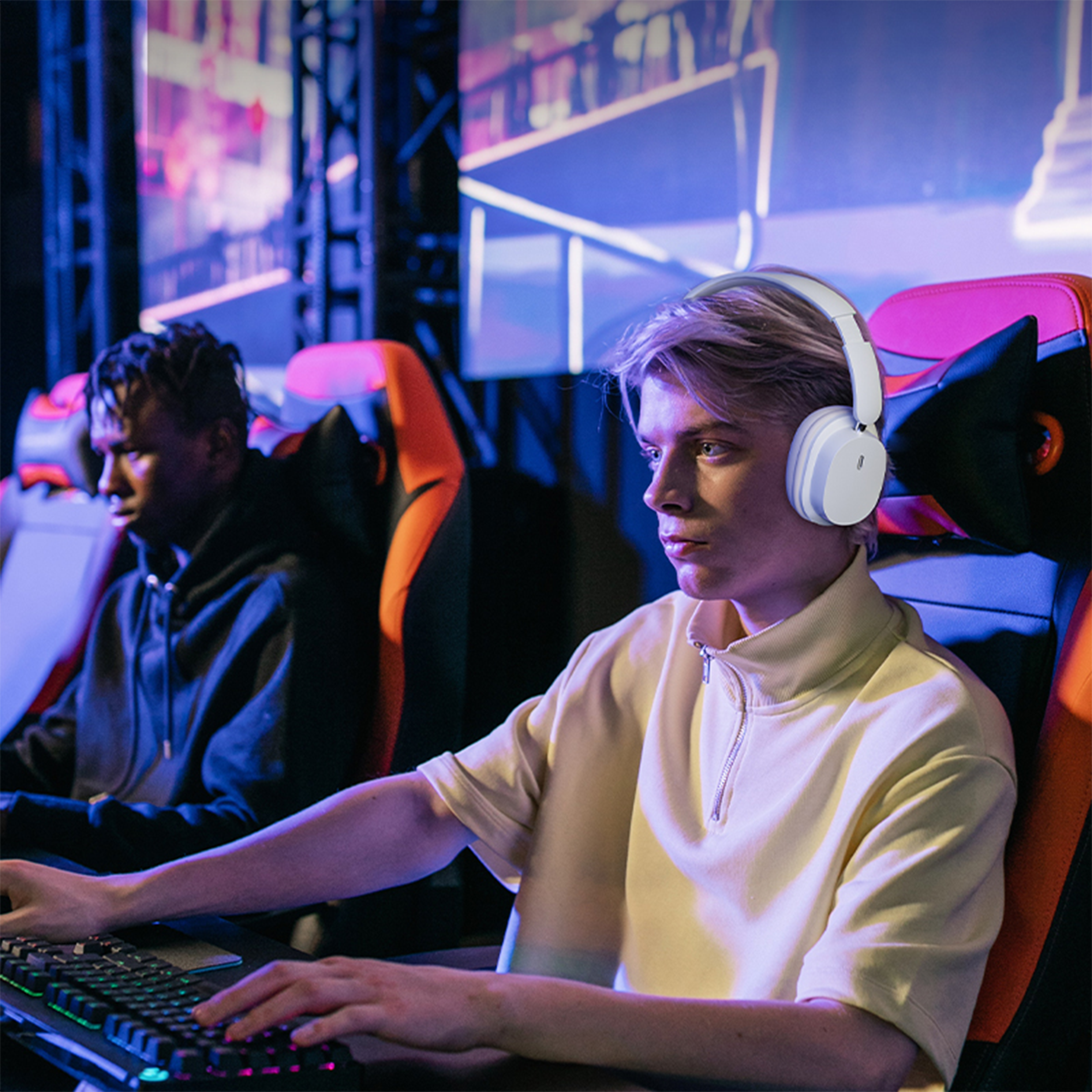 Zwei Spieler mit Kopfhörern, einer blond, spielen in einem Spielzimmer. Blaue und orangefarbene Lichter.