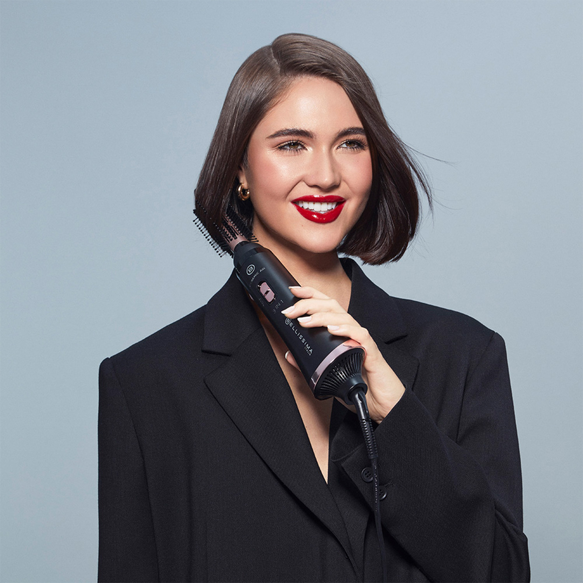 Frau hält Haarstyler. Lächelt, trägt schwarzen Blazer, roter Lippenstift.
