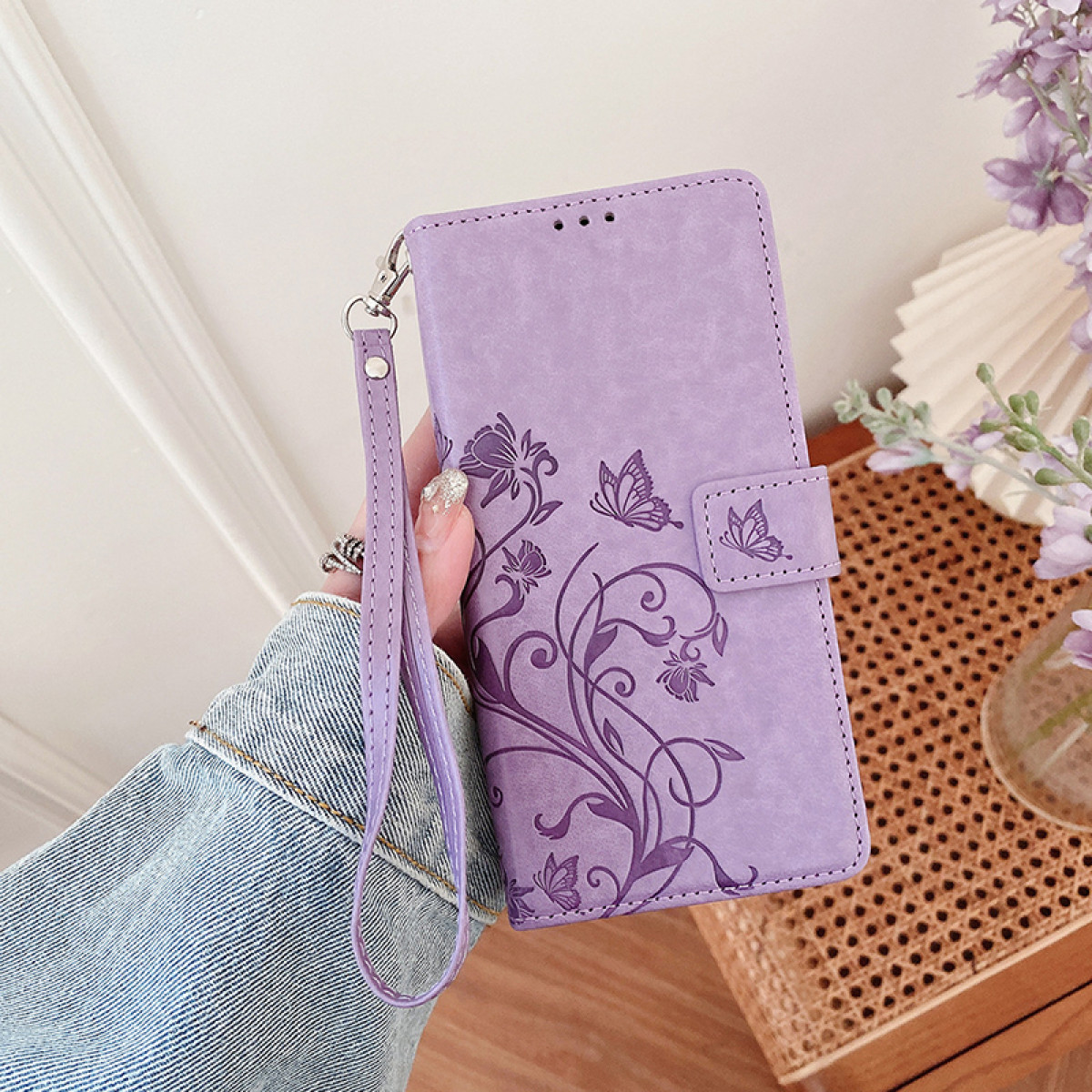 Custodia per telefono viola tenuta in mano. Ha un design a farfalla e un cinturino da polso. Una giacca di jeans è in vista.