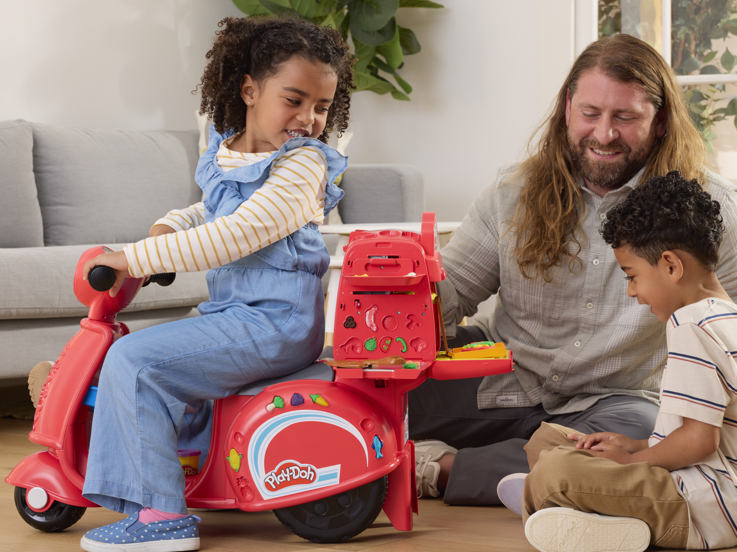 Niños y un adulto jugando con un scooter de juguete rojo con accesorios de comida.