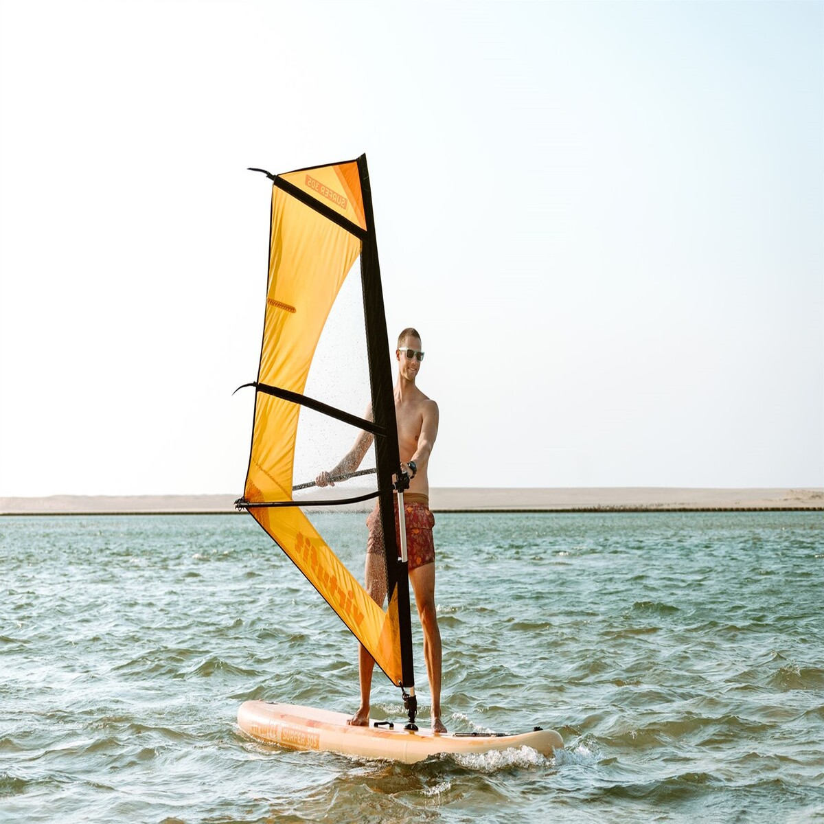 Ein Mann beim Windsurfen auf dem Wasser, hält ein Segel und steht auf einem Brett; heller Himmel.