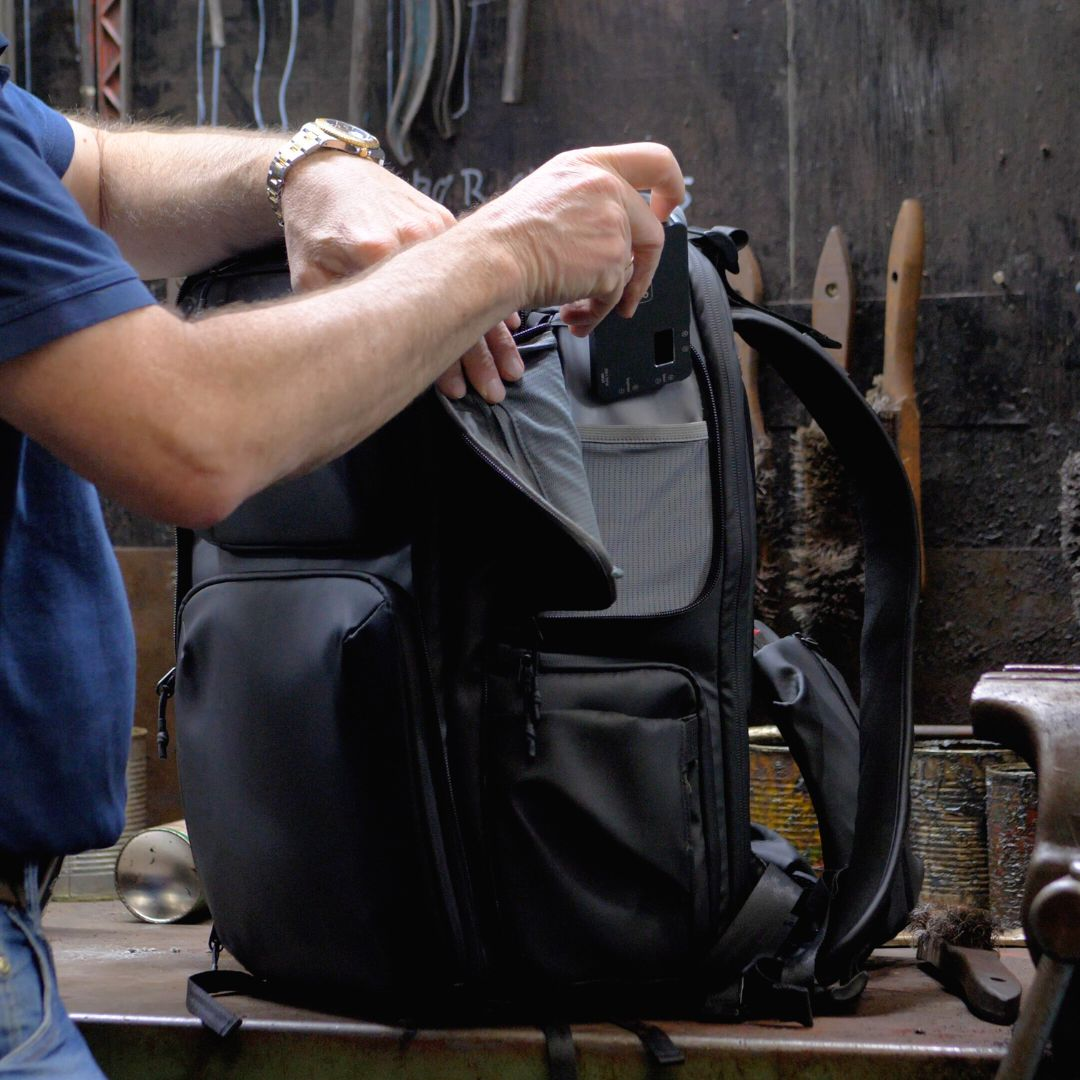 Man opening a black backpack in a workshop. Tools and supplies are in the background.