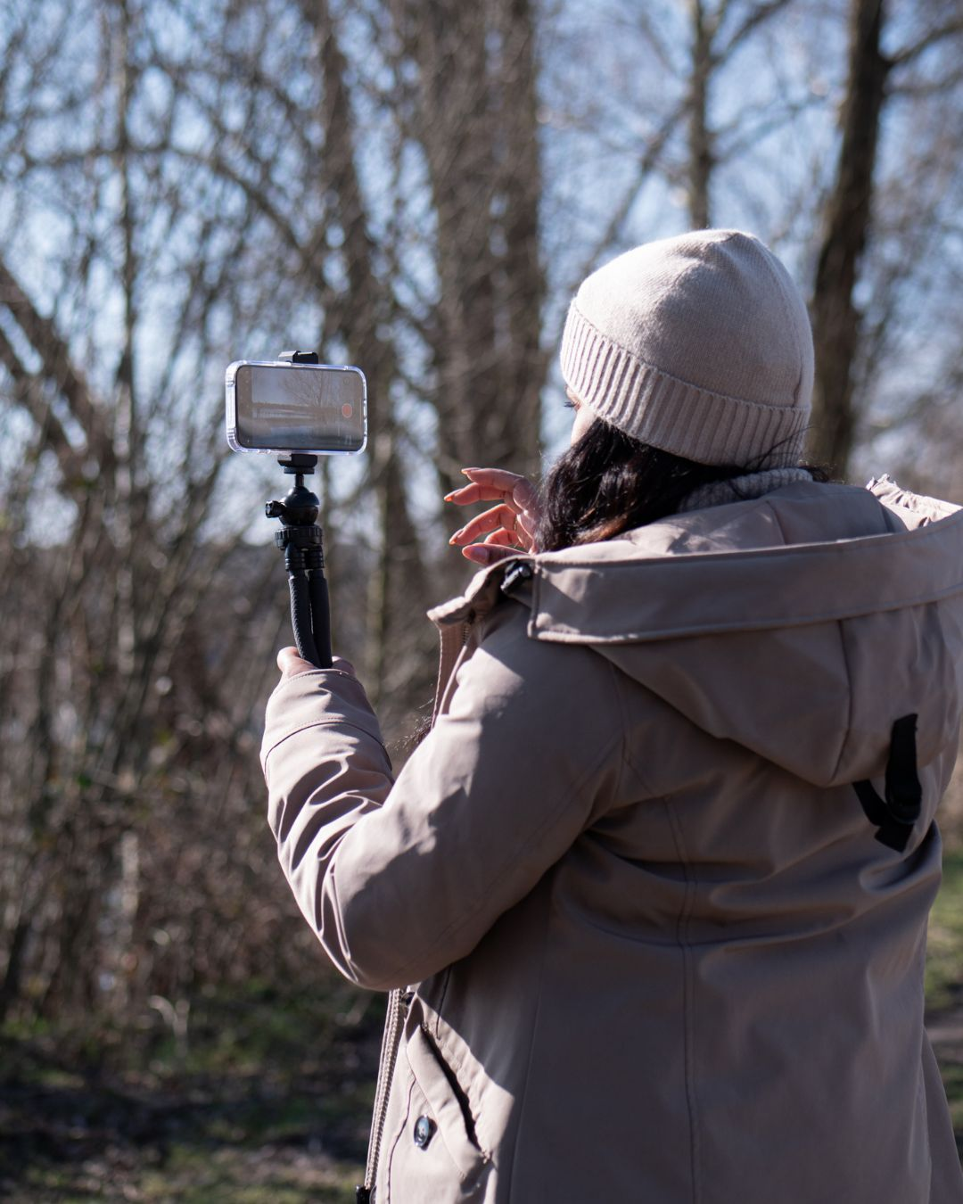 Frau filmt draußen mit Telefon und Selfie-Stick. Bäume im Hintergrund, sonniger Tag.