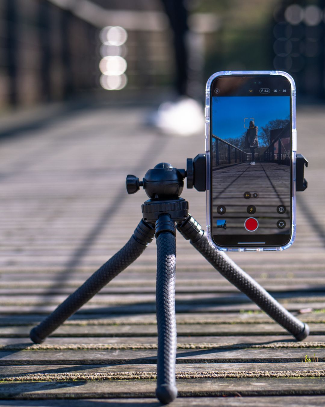 Ein Telefon auf einem Stativ. Der Bildschirm zeigt eine Person auf einer Brücke, mit Steuerelementen.