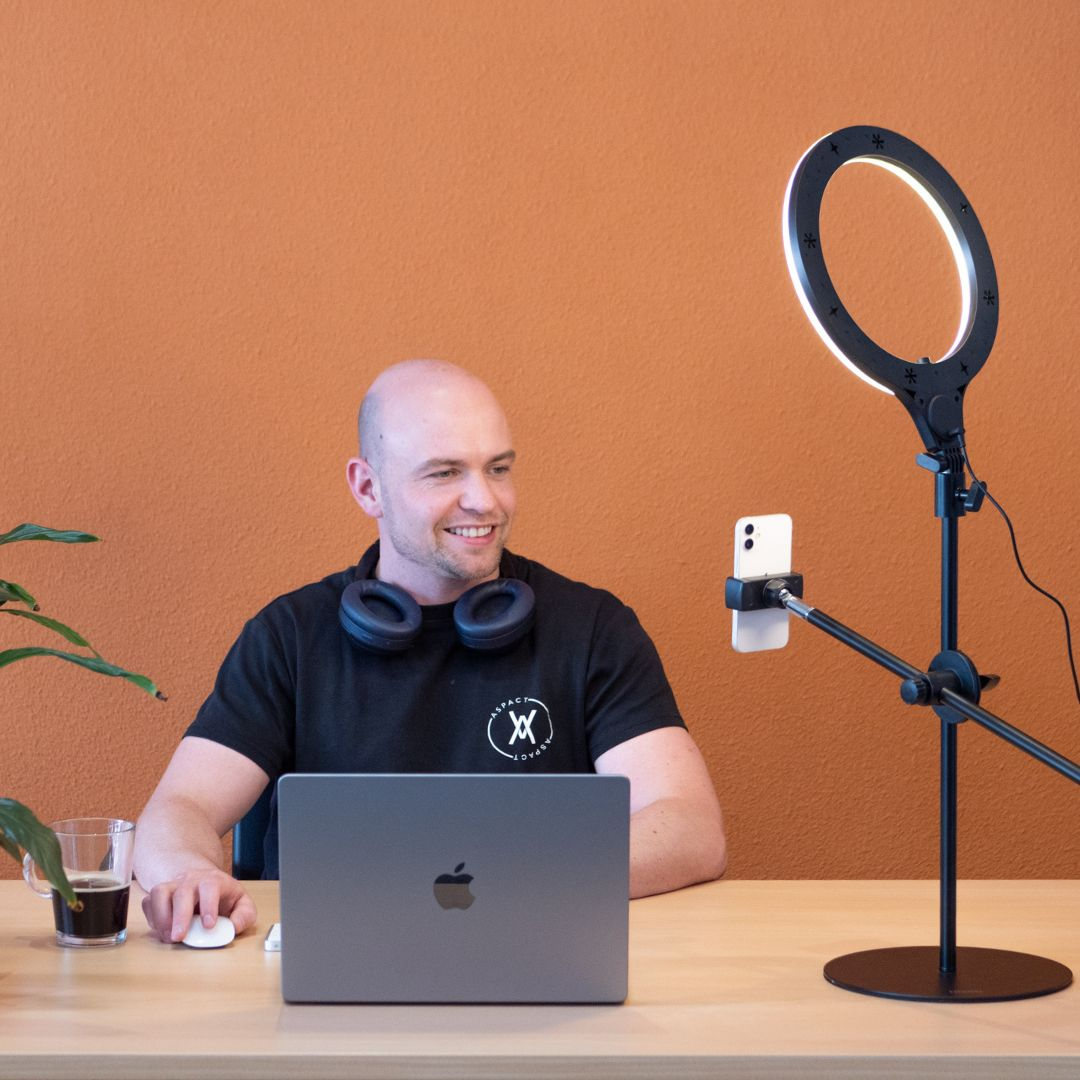 Een man werkt op een laptop met een ringlicht en telefoon, zittend aan een bureau.