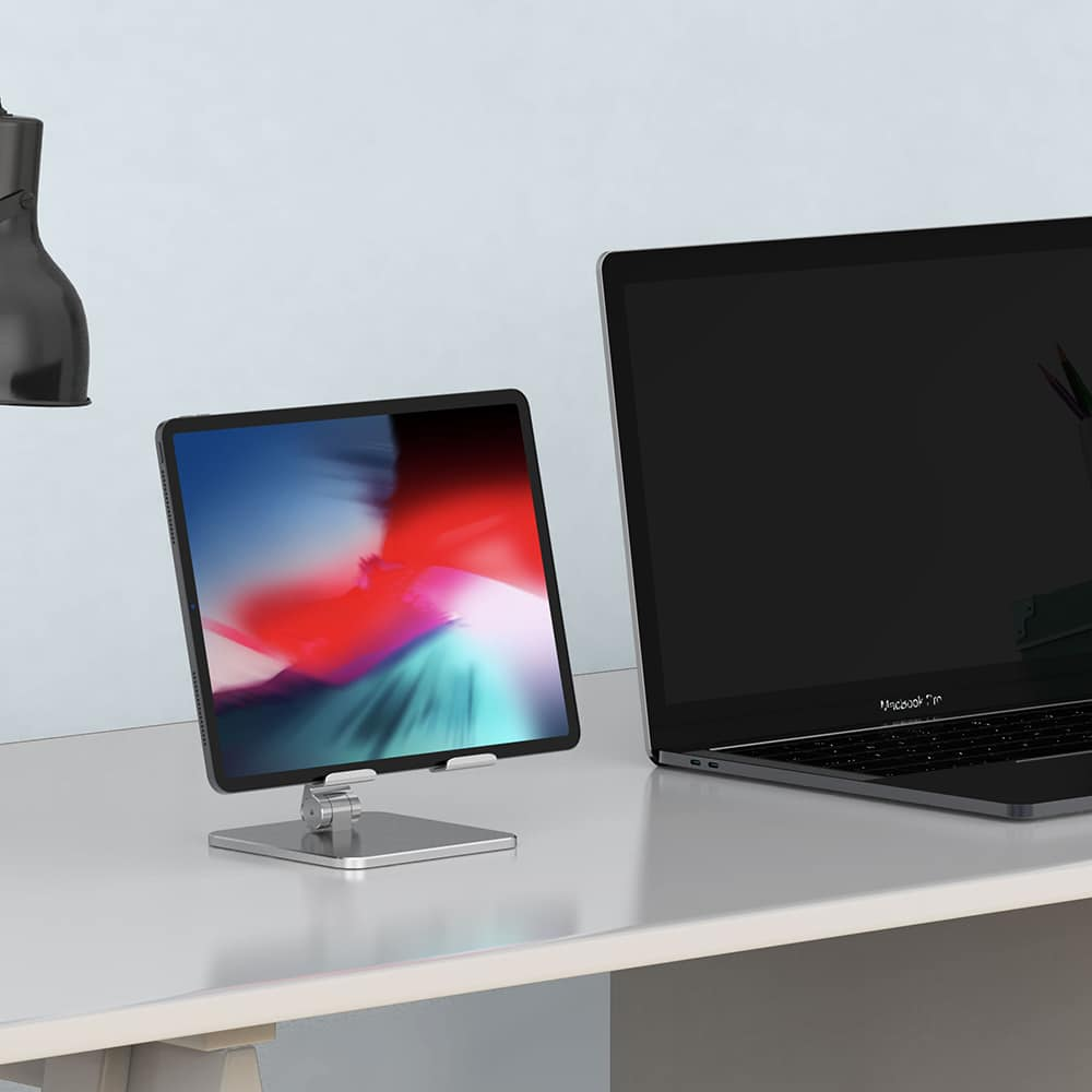Une tablette sur un support à côté d'un ordinateur portable. Les deux sont sur un bureau blanc. Lampe noire en arrière-plan.