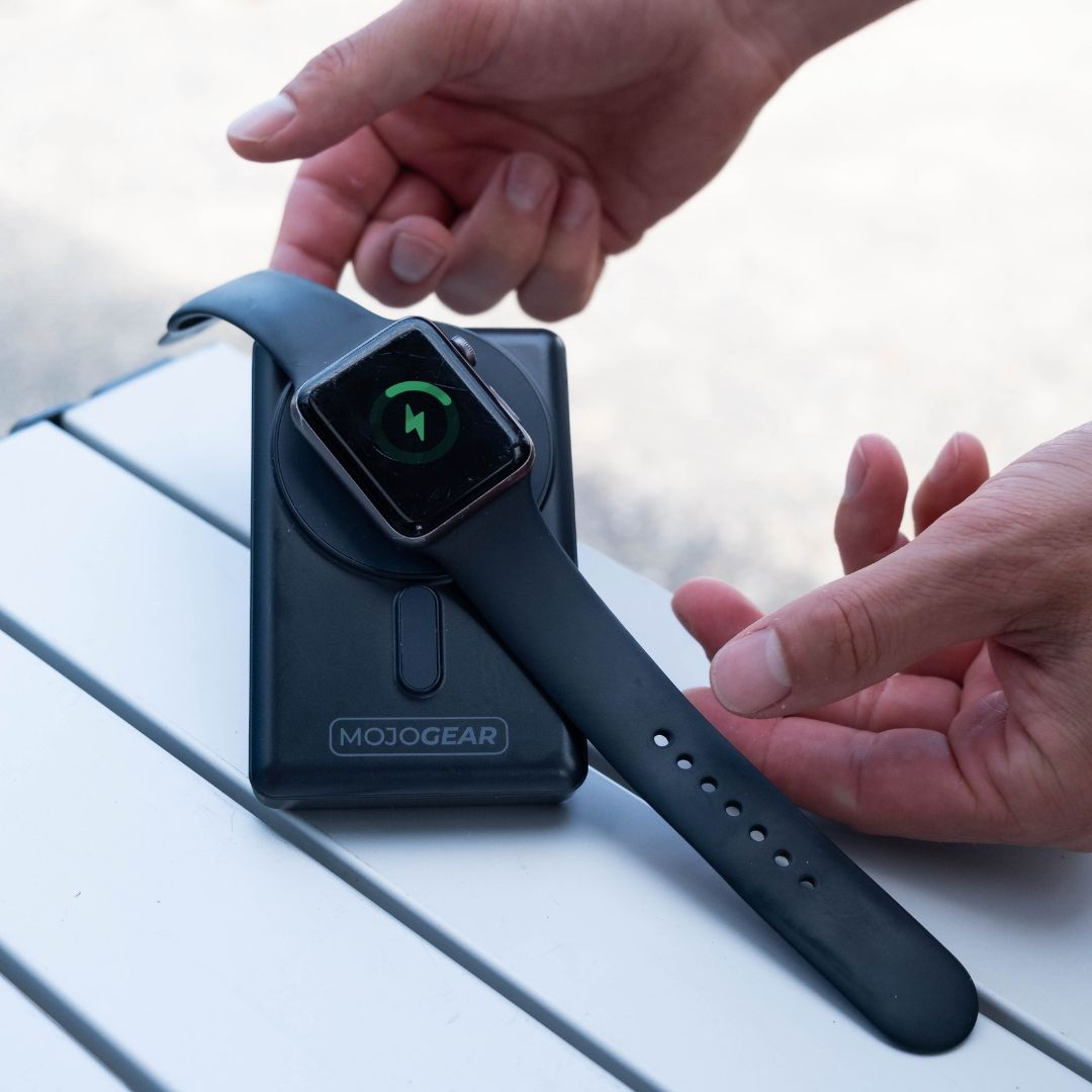 A person's hands holding a black smartwatch on a charging station. White table background.
