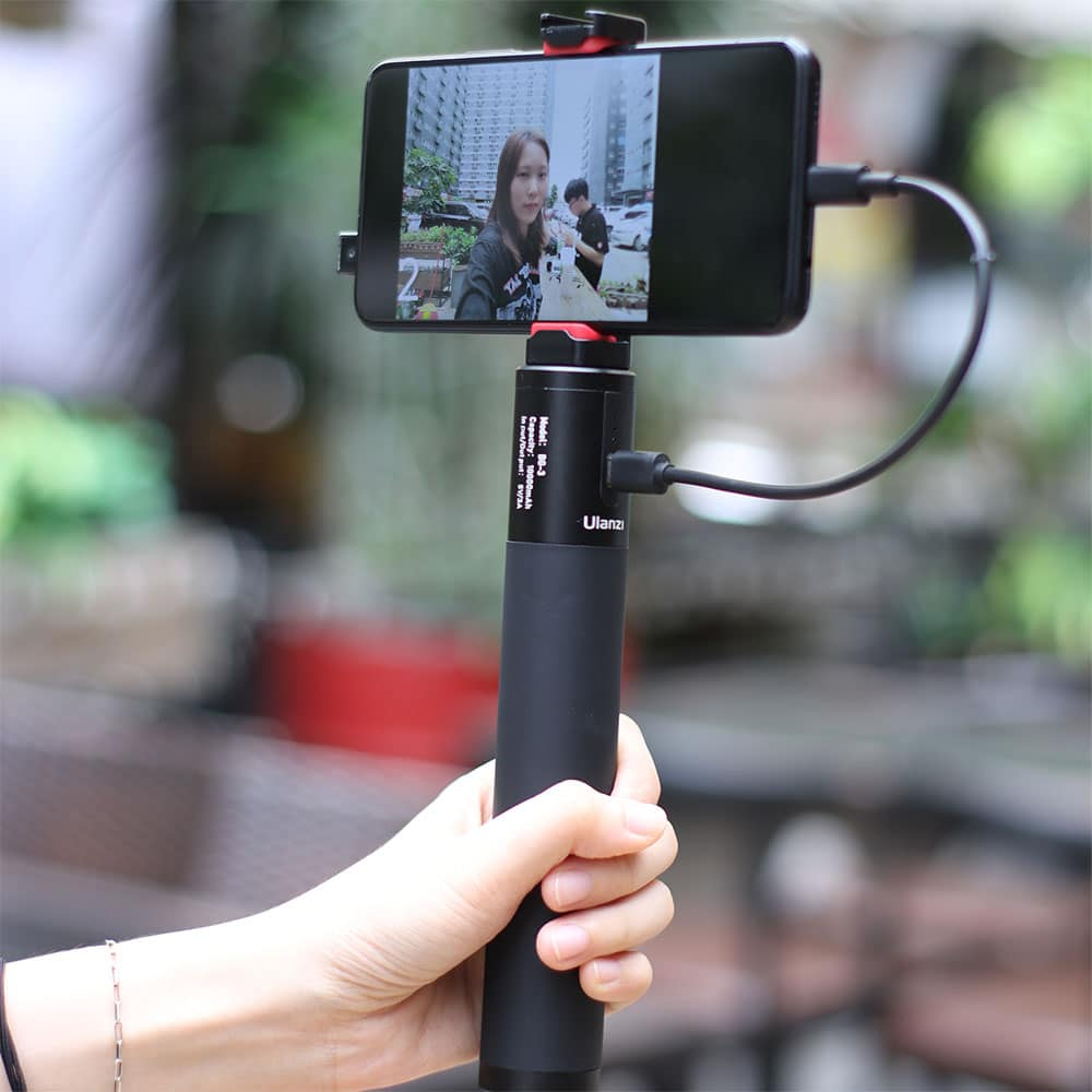 Een hand die een zwarte selfiestick vasthoudt met een telefoon die een videogesprek toont.