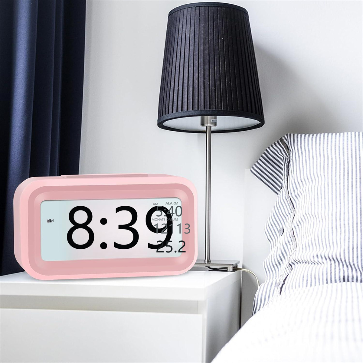 Pinker digitaler Wecker auf einem weißen Nachttisch, neben einer Lampe. Bett und Wand im Hintergrund.