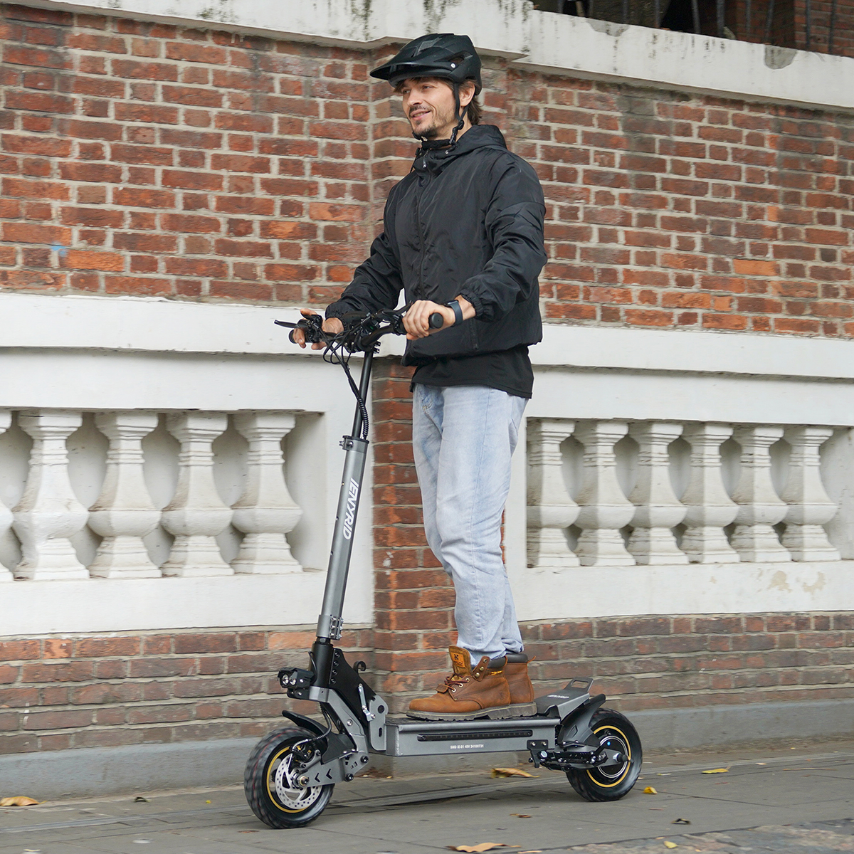 Mężczyzna jedzie szarą hulajnogą elektryczną po chodniku. Ma kask i kurtkę. Ceglany mur w tle.