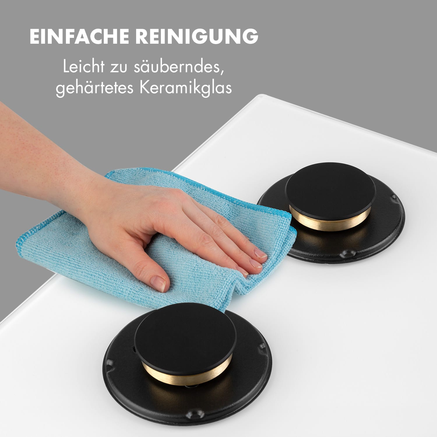 Eine Hand wischt eine weiße Kochfeldplatte ab. Zwei schwarze Herdplatten mit goldenem Ring sind sichtbar.