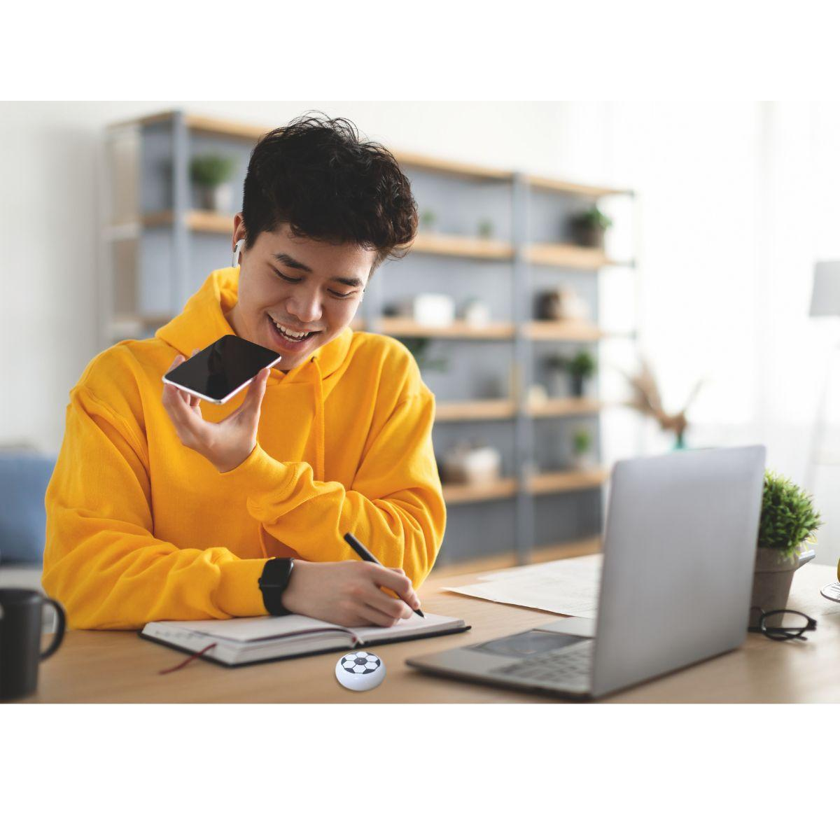 Ein Mann in einem gelben Kapuzenpullover telefoniert, während er in ein Notizbuch schreibt, mit einem Laptop.