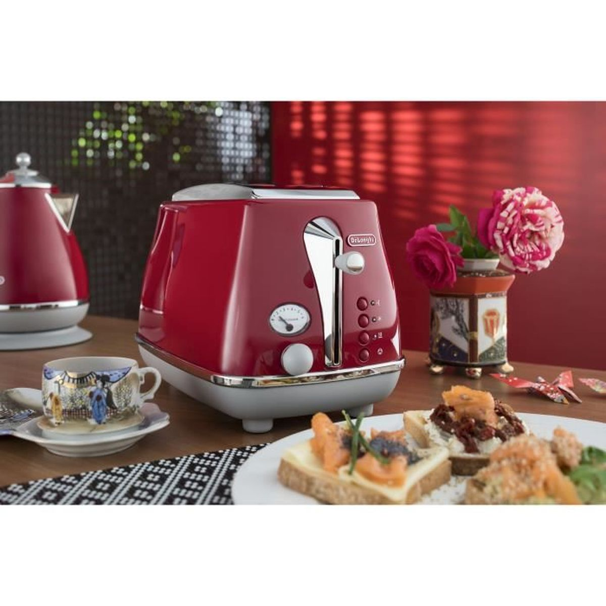 Ein roter Toaster mit einem Teller Sandwiches und anderem Essen auf einem Holztisch. Blumen im Hintergrund.