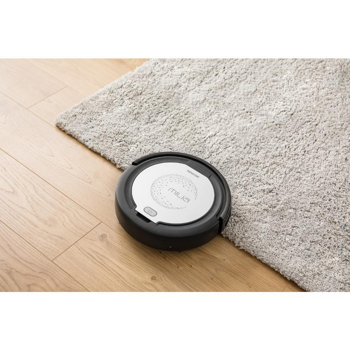 Aspirateur robot noir et argenté sur tapis gris clair et sol en bois.