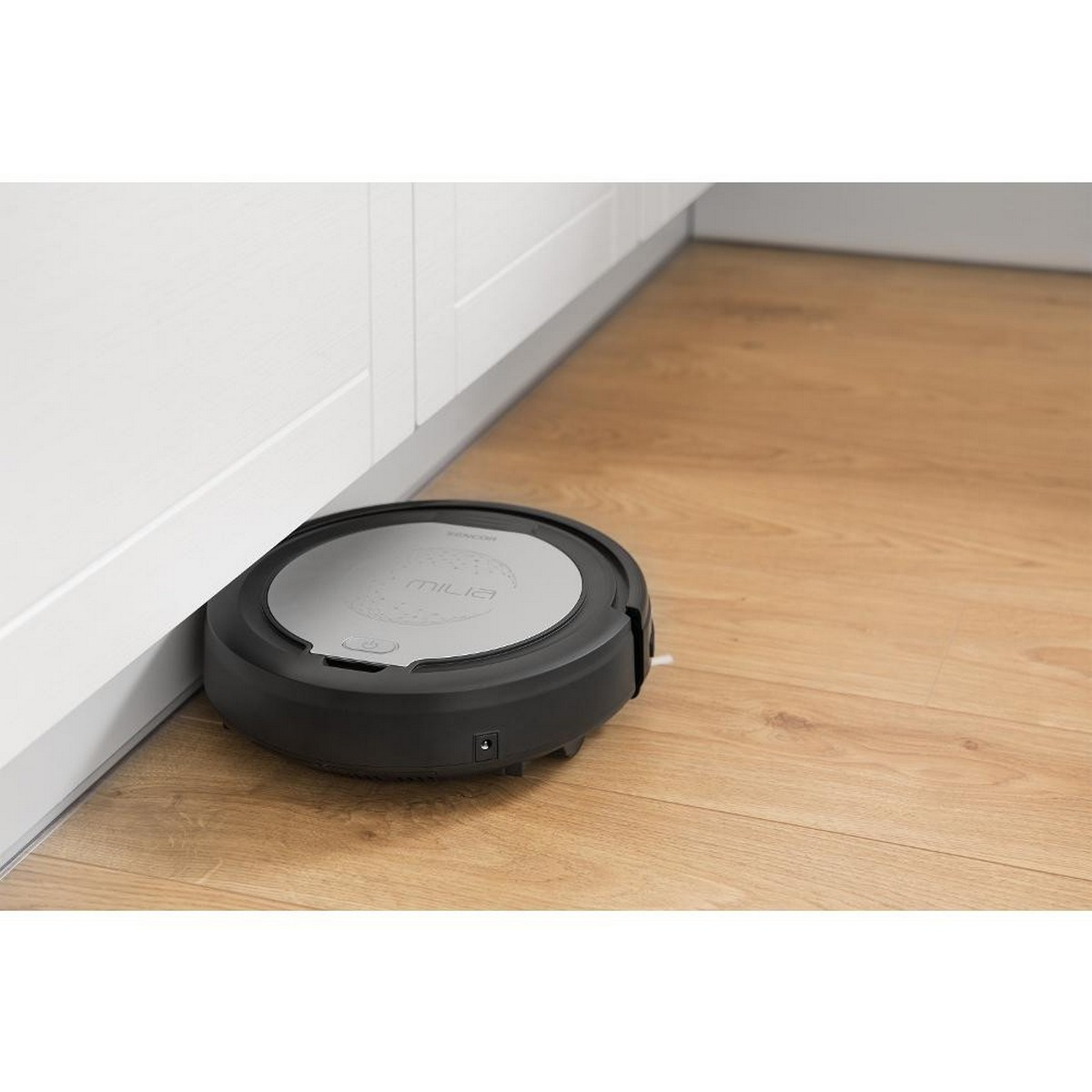 Un aspirateur robot noir et gris sur un plancher en bois près d'un meuble blanc.