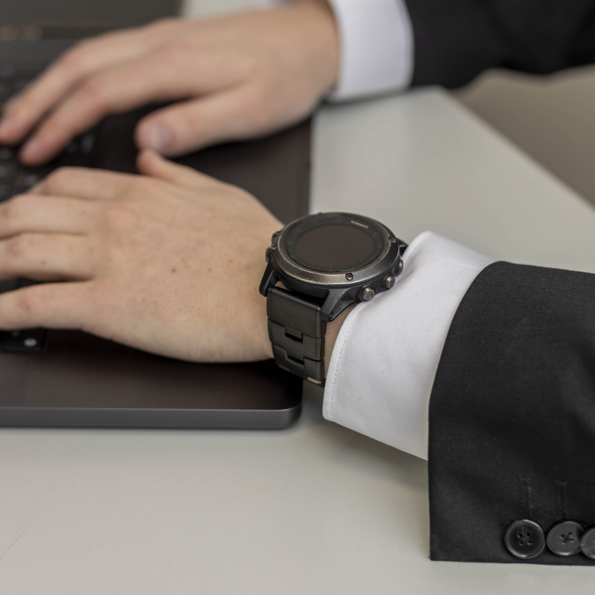 Eine Person, die eine schwarze Uhr am Handgelenk trägt, tippt auf einem Laptop auf einem weißen Schreibtisch.