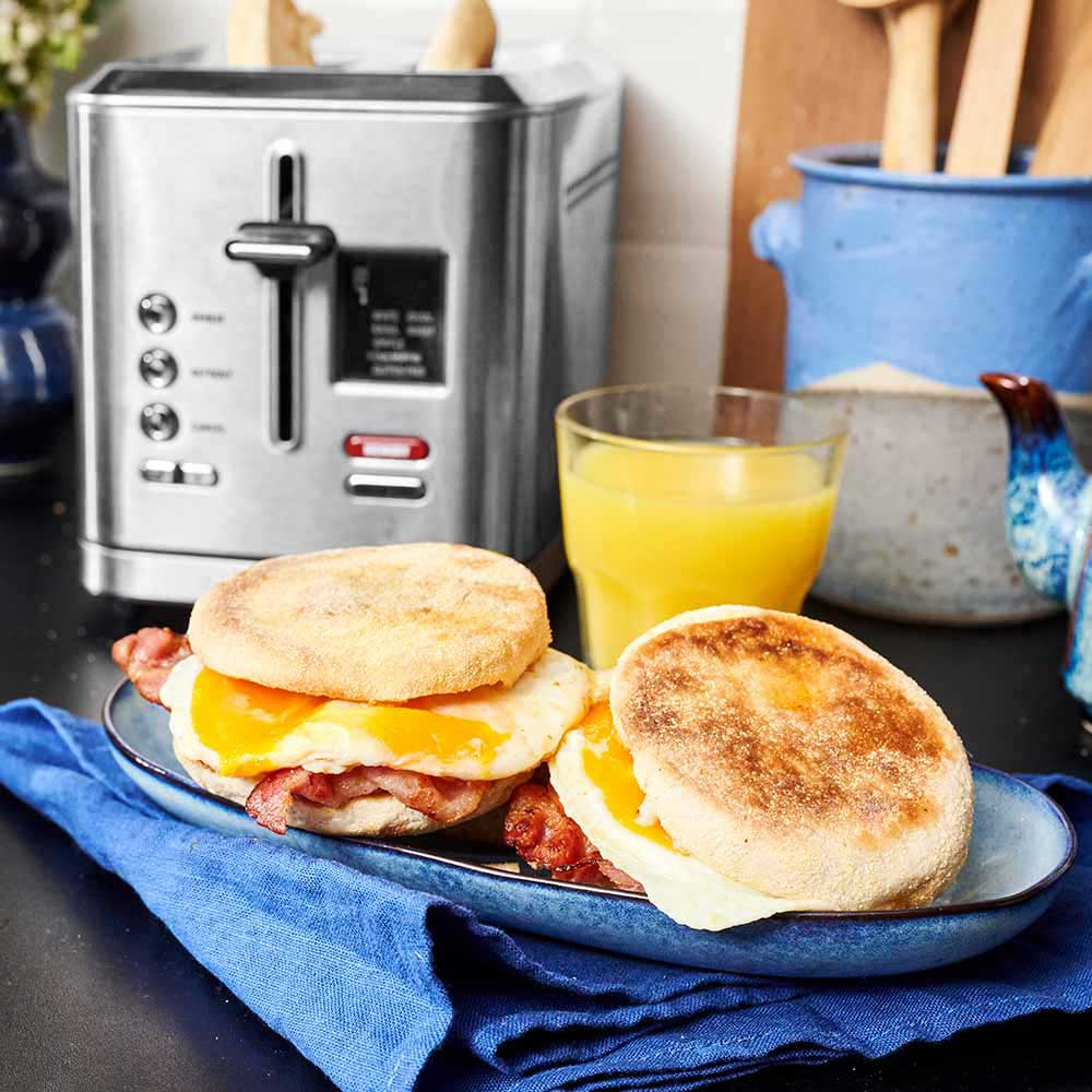 Frühstück mit englischen Muffins, Speck und Spiegeleiern. Toaster, Saft und Utensilien. Küchenumgebung.
