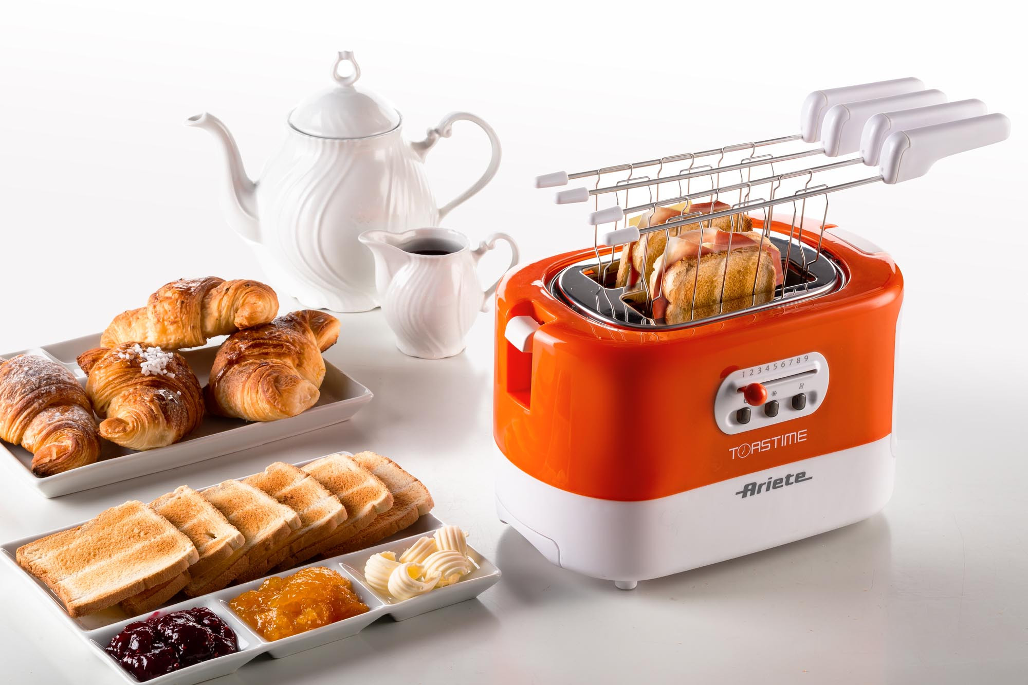 Oranje en witte broodrooster met toast, croissants en kruiderijen op een witte tafel.