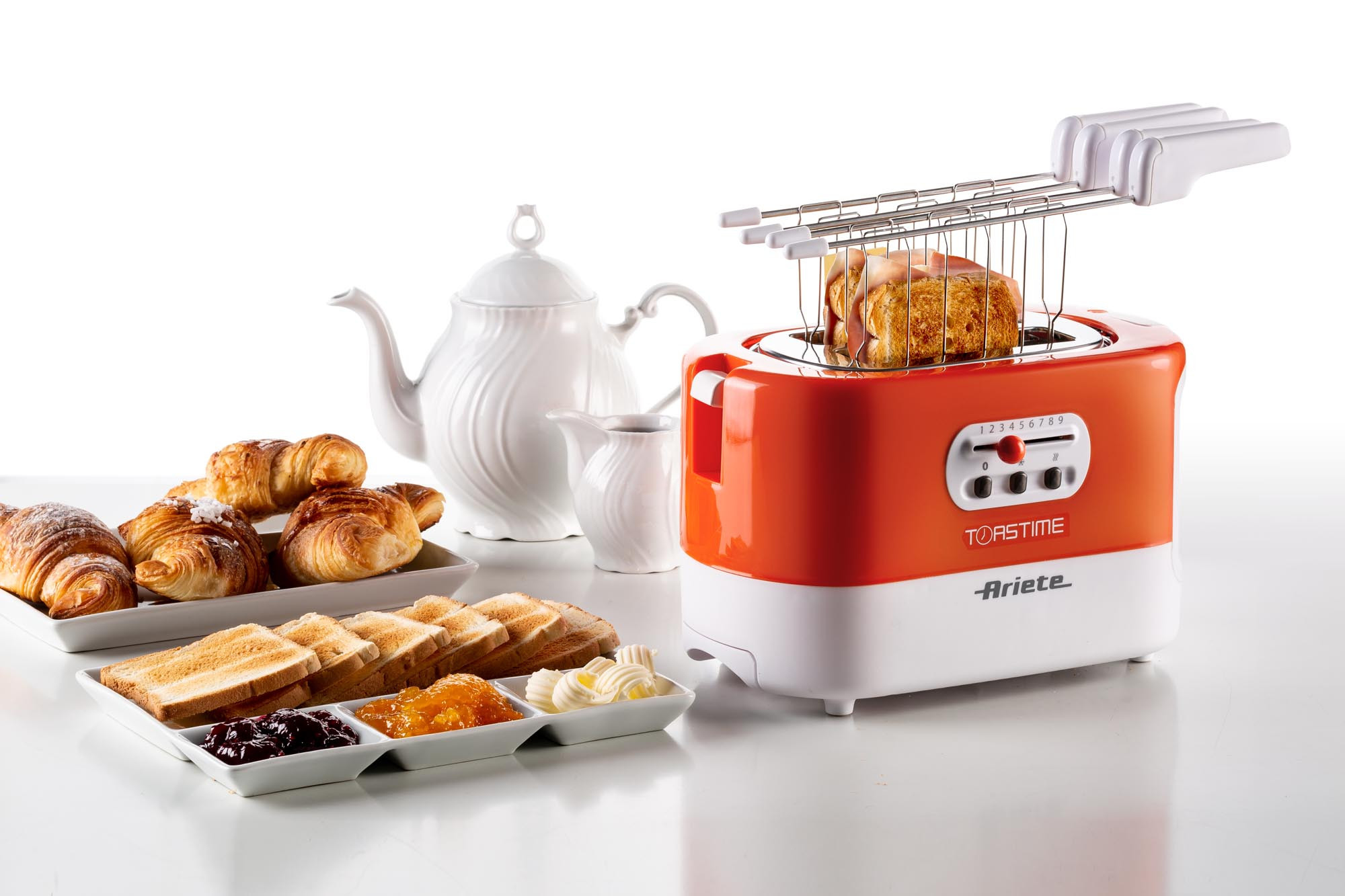 Broodrooster met geroosterd brood, croissants en jam op een wit oppervlak. Theepot en boter zijn ook aanwezig.
