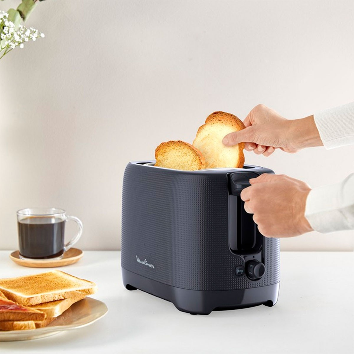 Een persoon die brood roostert in een donkergrijze broodrooster met een kopje koffie en andere toast.