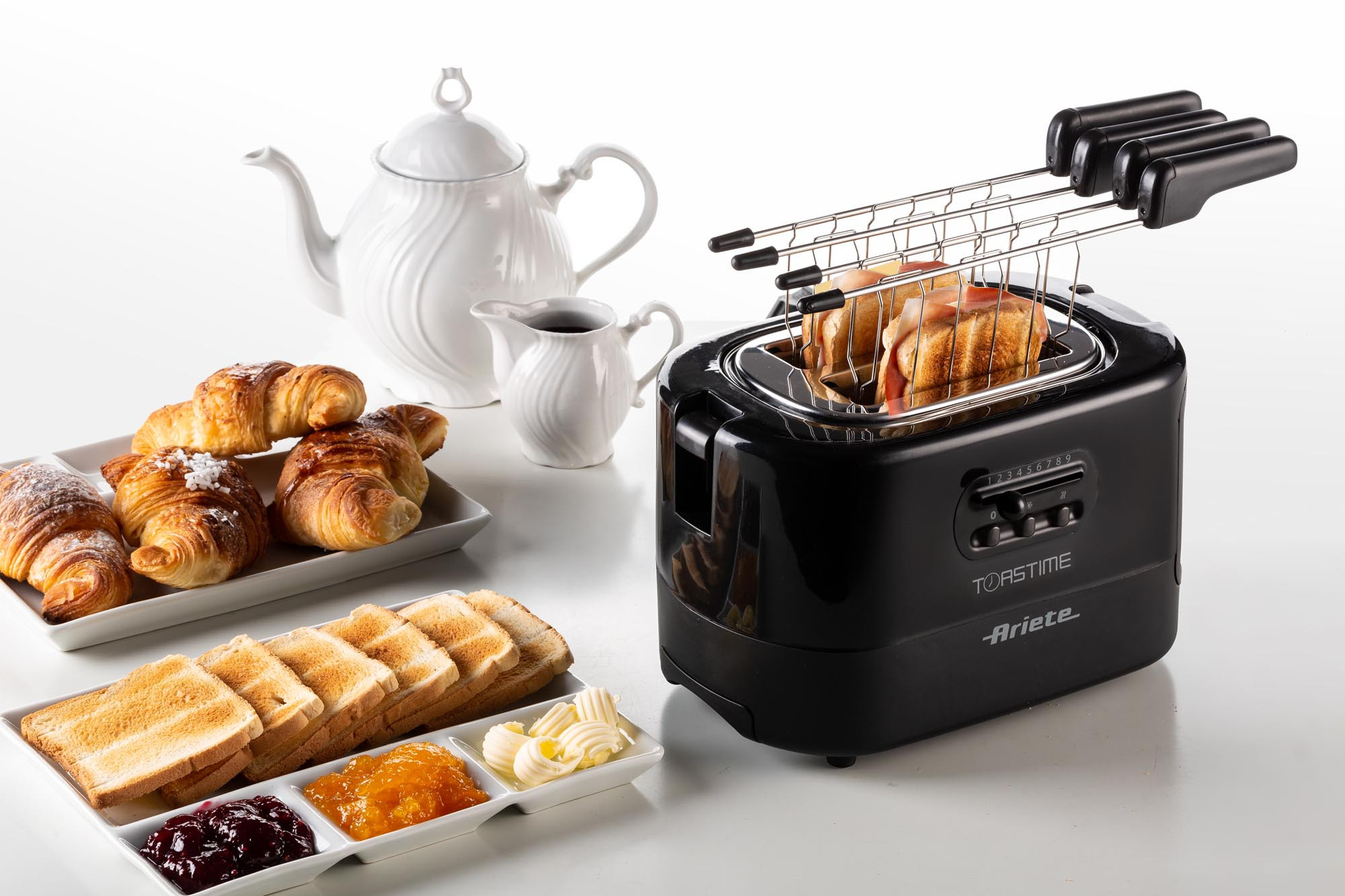 Een zwarte broodrooster met toast, croissants en kruiderijen op een wit oppervlak, ontbijtscène.