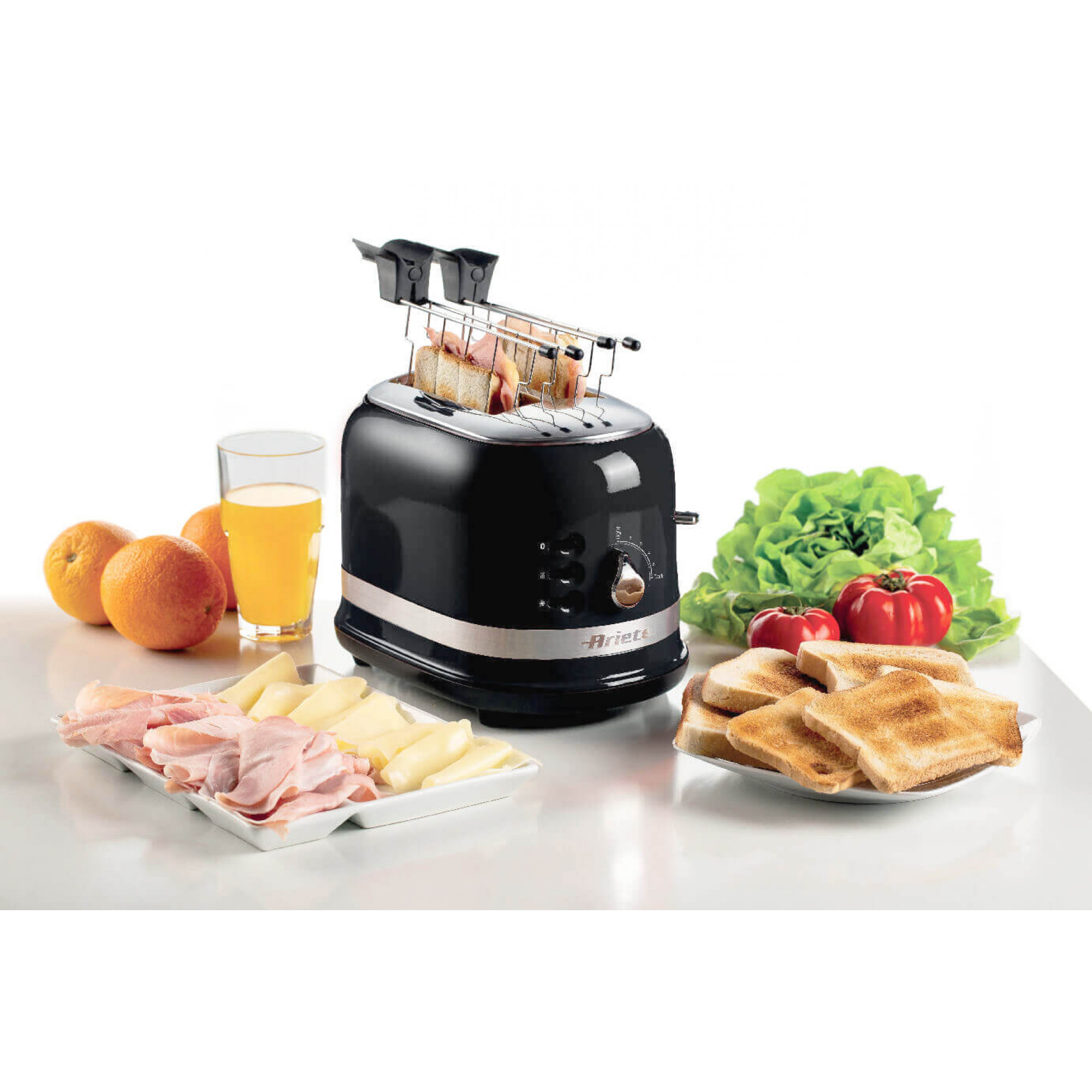 Een broodrooster met toast, brood, ham en sinaasappelsap op een witte tafel. Groene sla aan de zijkant.