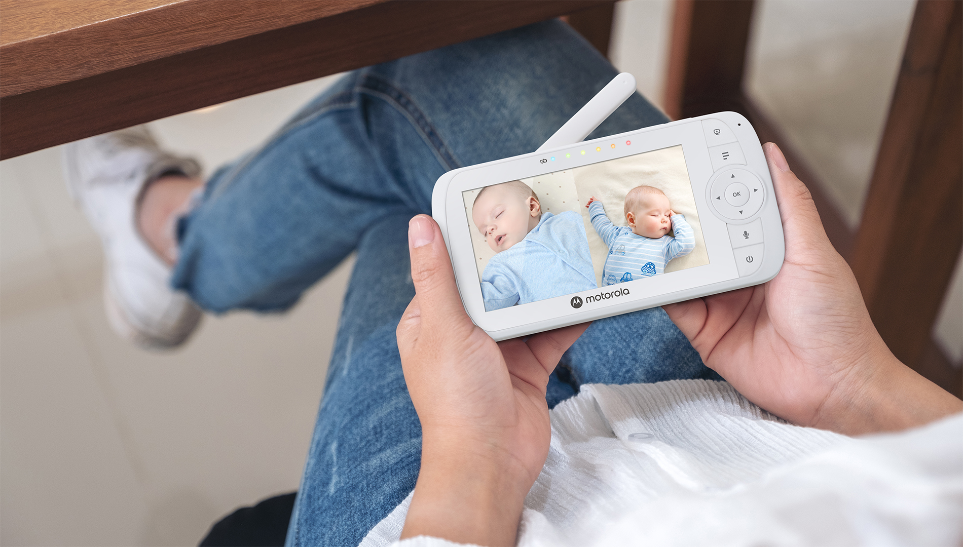 Hands holding a Motorola baby monitor. Screen shows two babies sleeping. Wooden table background.