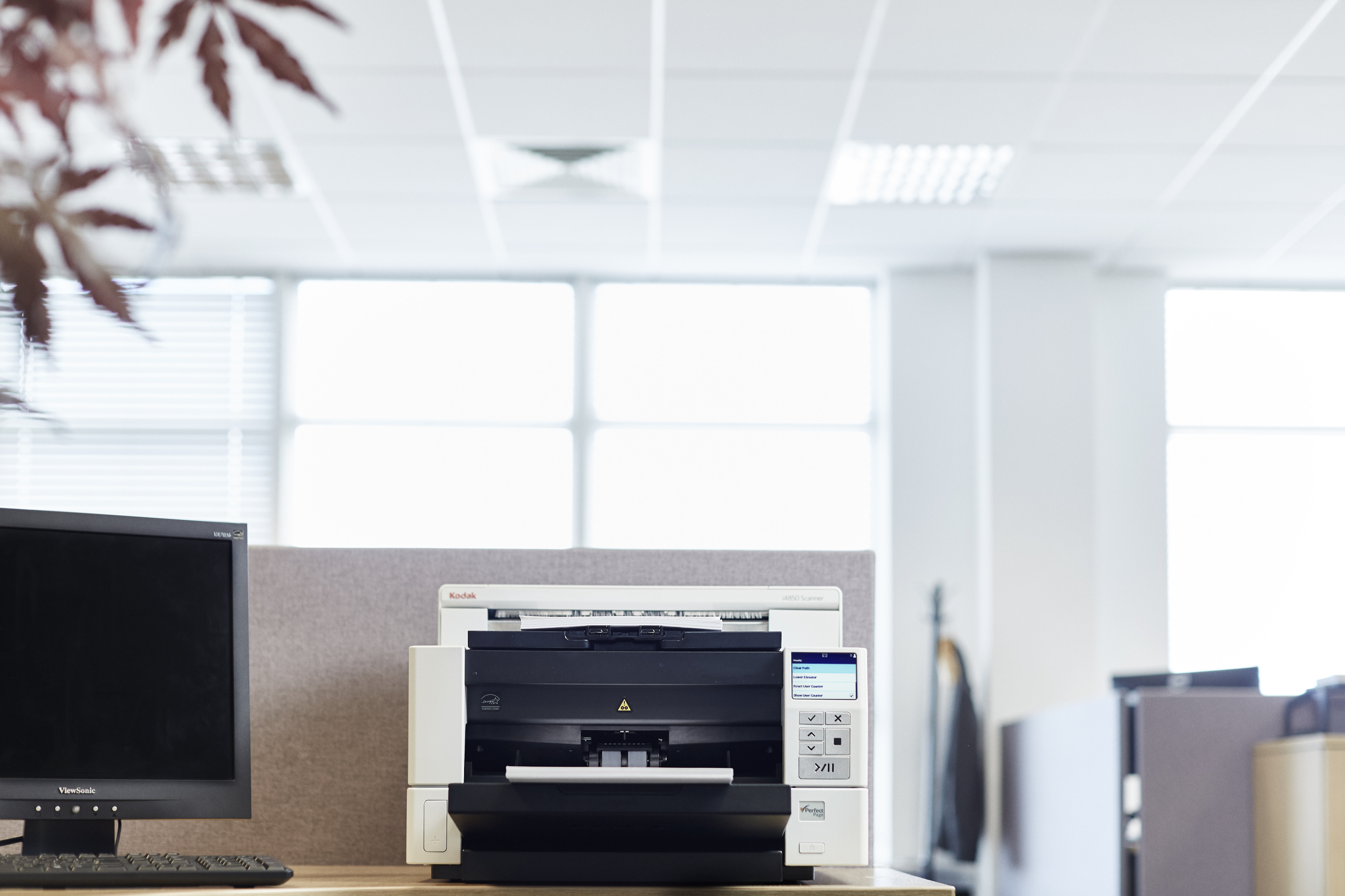Ein Bürotisch mit Computermonitor, Drucker und Fenster im Hintergrund.