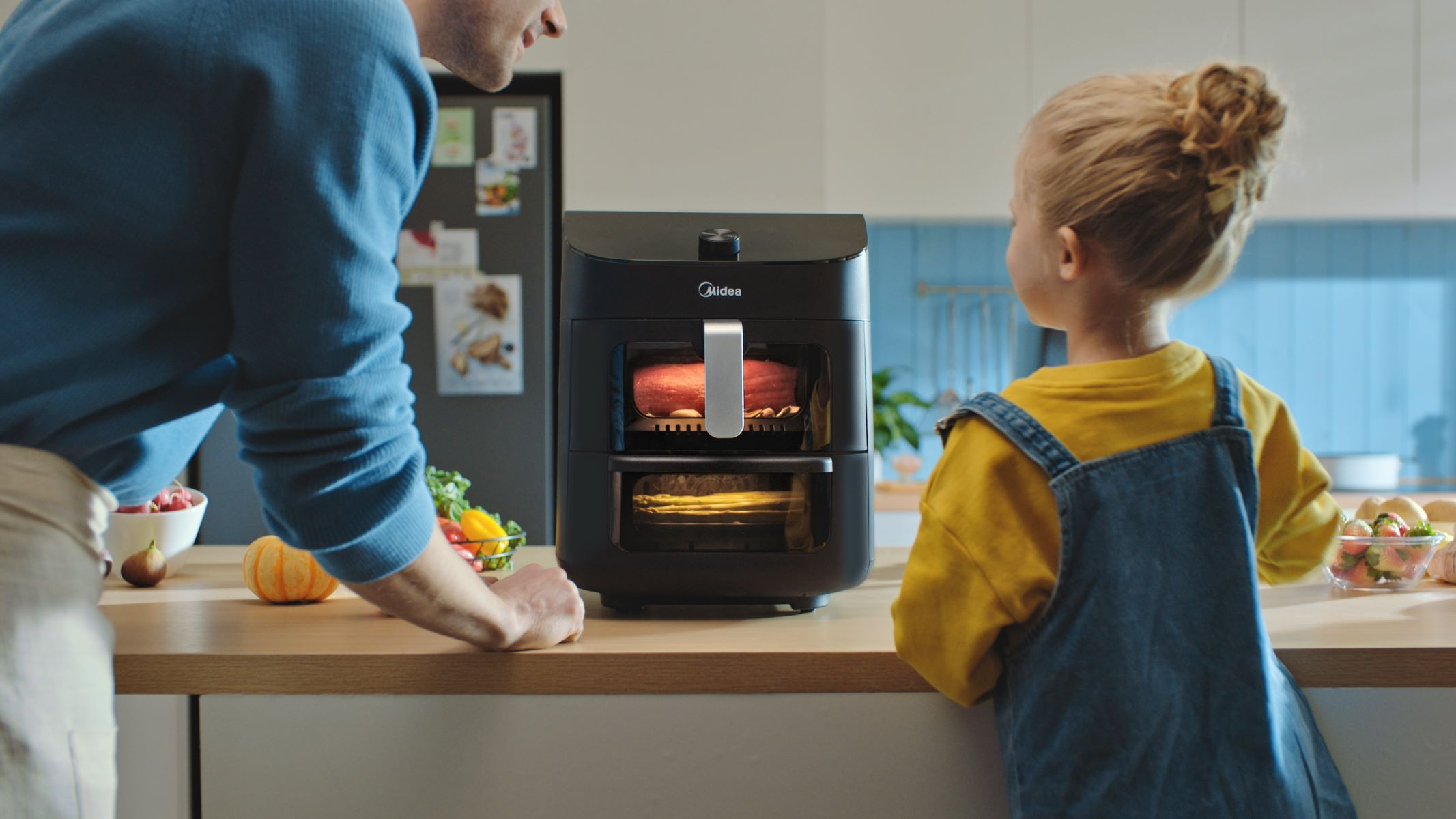 Man en kind bij een zwarte airfryer met eten. Toonbank met groenten.