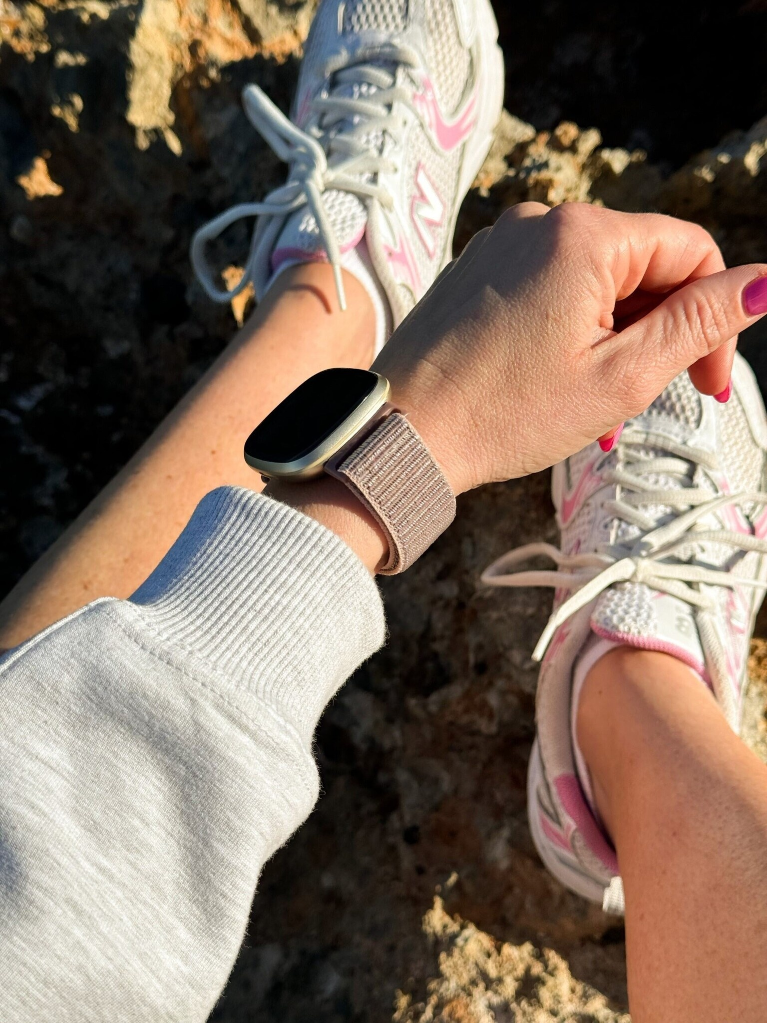 Hand mit Uhr, pinken Nägeln, weißen Schuhen und heller Kleidung auf Stein.